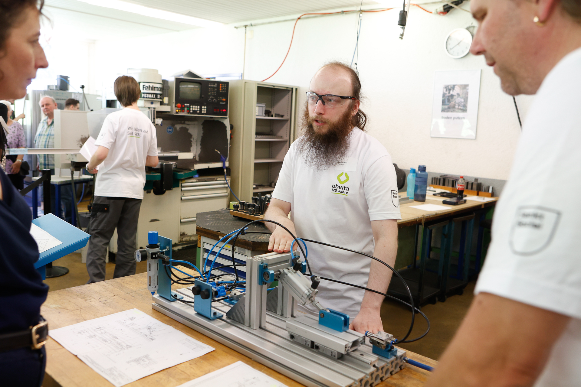 Ein Mann, der eine Schutzbrille und ein weißes T-Shirt trägt, bespricht mit zwei Kollegen in einer Werkstatt eine mechanische Vorrichtung. Auf dem Tisch liegen technische Zeichnungen, und im Hintergrund sind weitere Personen bei der Arbeit zu sehen.