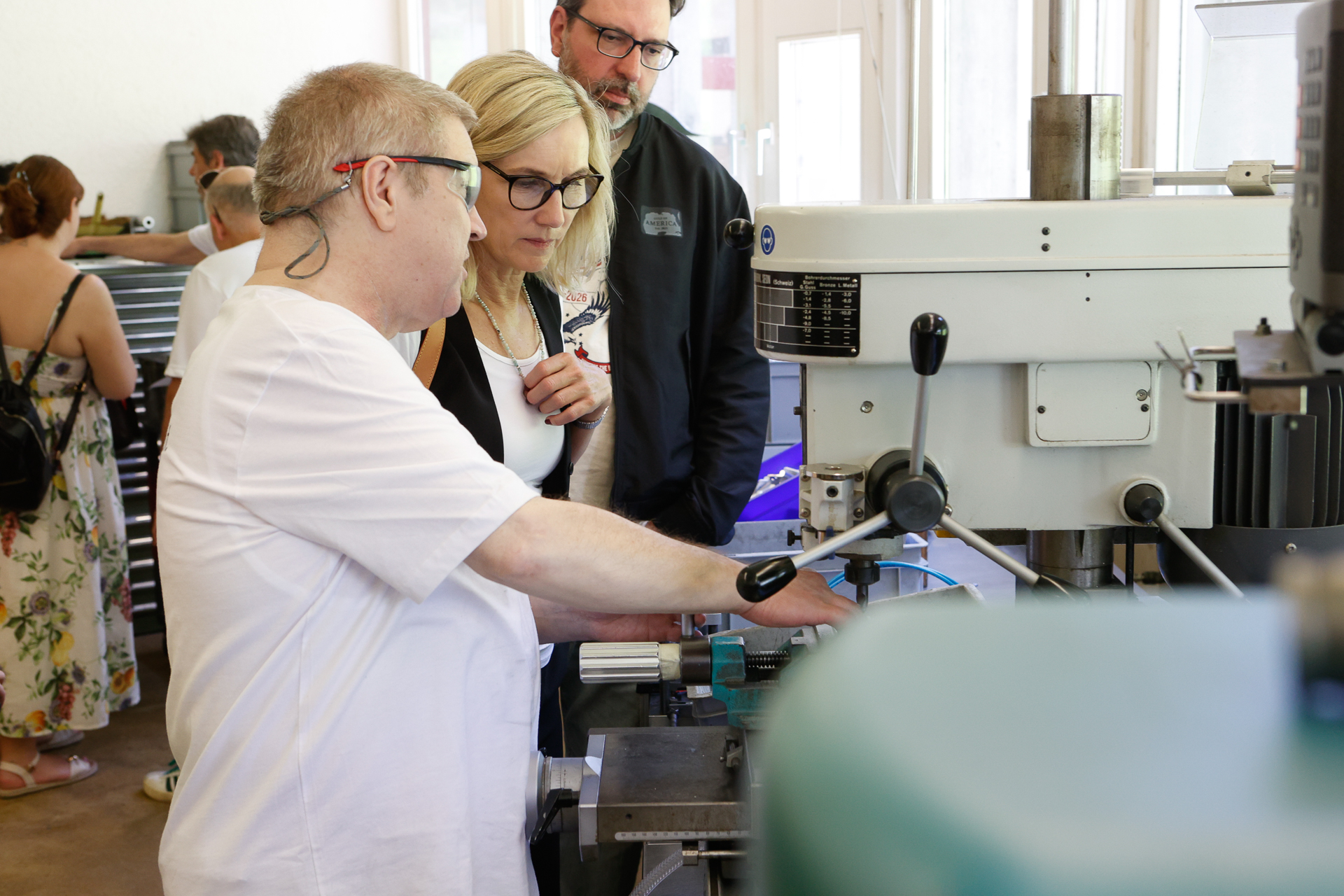 Ein Mann mit Schutzbrille zeigt zwei Zuschauern in einer Werkstatt, wie man eine Werkzeugmaschine bedient. Im Hintergrund sind weitere Personen zu sehen, die verschiedenen Tätigkeiten nachgehen.