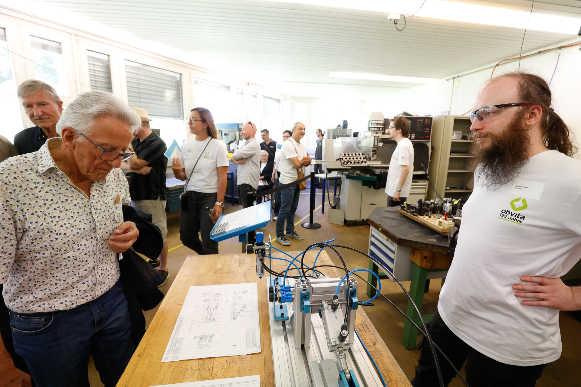 In einer Werkstatt versammelt sich eine Gruppe von Menschen um einen Tisch, auf dem technische Zeichnungen und Maschinen liegen. Ein Mann mit Schutzbrille erklärt einem älteren Mann etwas, während andere im Hintergrund zuschauen.
