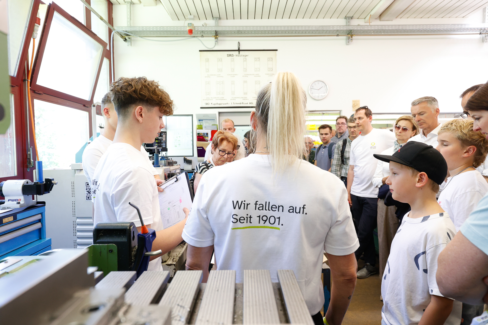 Eine Gruppe von Menschen in einem Workshop hört einem jungen Referenten zu. Die meisten tragen weiße T-Shirts; auf einem steht: „Wir fallen auf. Seit 1901.“ Rundherum stehen Werkzeuge und Maschinen, und durch die Fenster fällt Tageslicht herein.