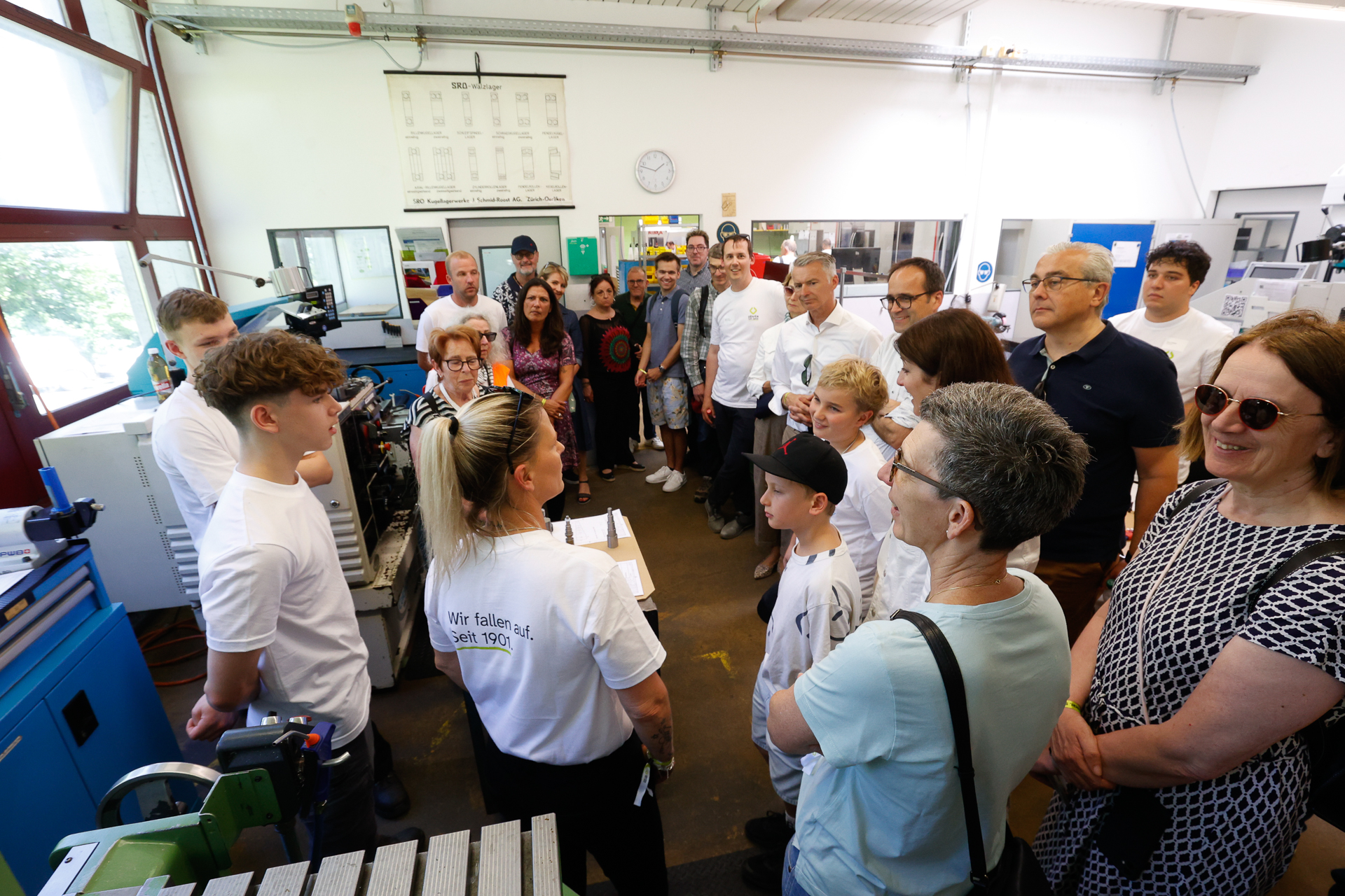 Eine Gruppe aus Erwachsenen und Kindern steht in einem Workshop und hört zwei jungen Leuten in weißen Hemden zu, die eine Vorführung geben. Man sieht Maschinen und Werkzeuge, und alle wirken interessiert und aufmerksam.