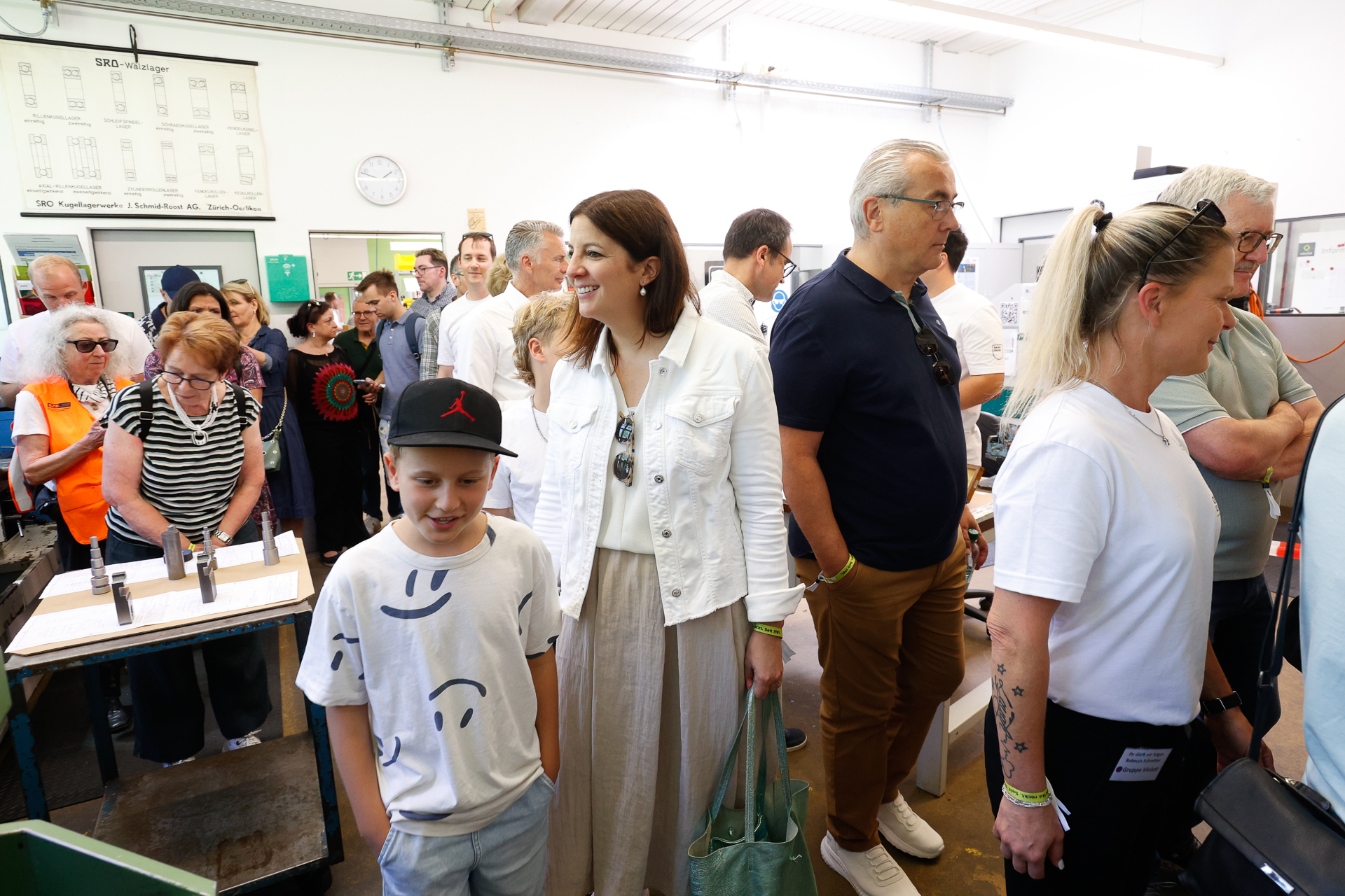 Eine Gruppe von Menschen, darunter Erwachsene und Kinder, steht in einem Innenraum dicht beieinander, lächelt und unterhält sich. Der Schauplatz scheint eine Werkstatt oder ein Industriebereich zu sein, im Hintergrund sind Werkzeuge und Maschinen zu sehen.