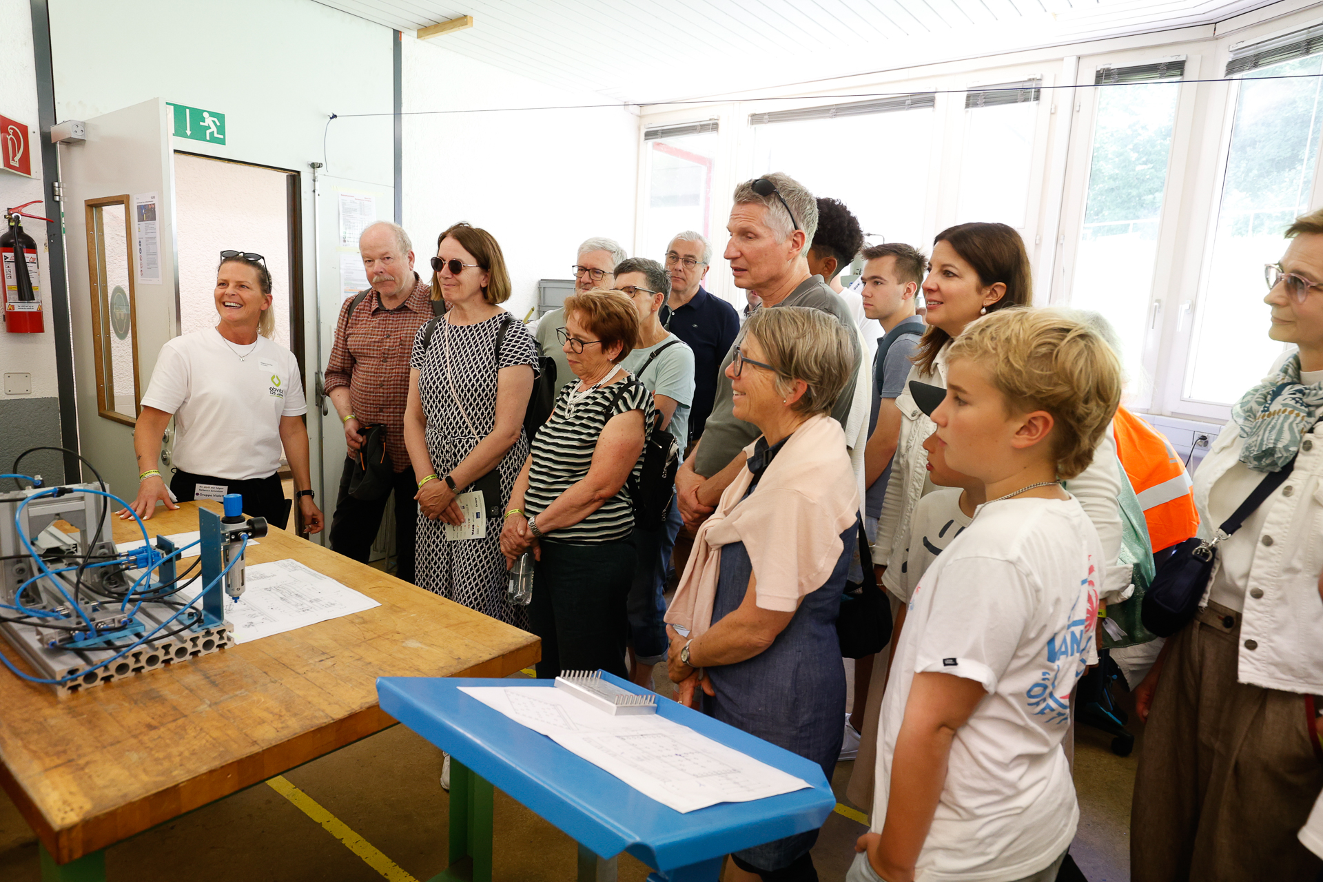 Eine Gruppe von Menschen steht in einer Werkstatt und verfolgt aufmerksam die Vorführung einer lächelnden Frau, die neben einer Werkbank mit Bauplänen und Maschinen steht. Im Hintergrund strömt Sonnenlicht durch große Fenster herein.