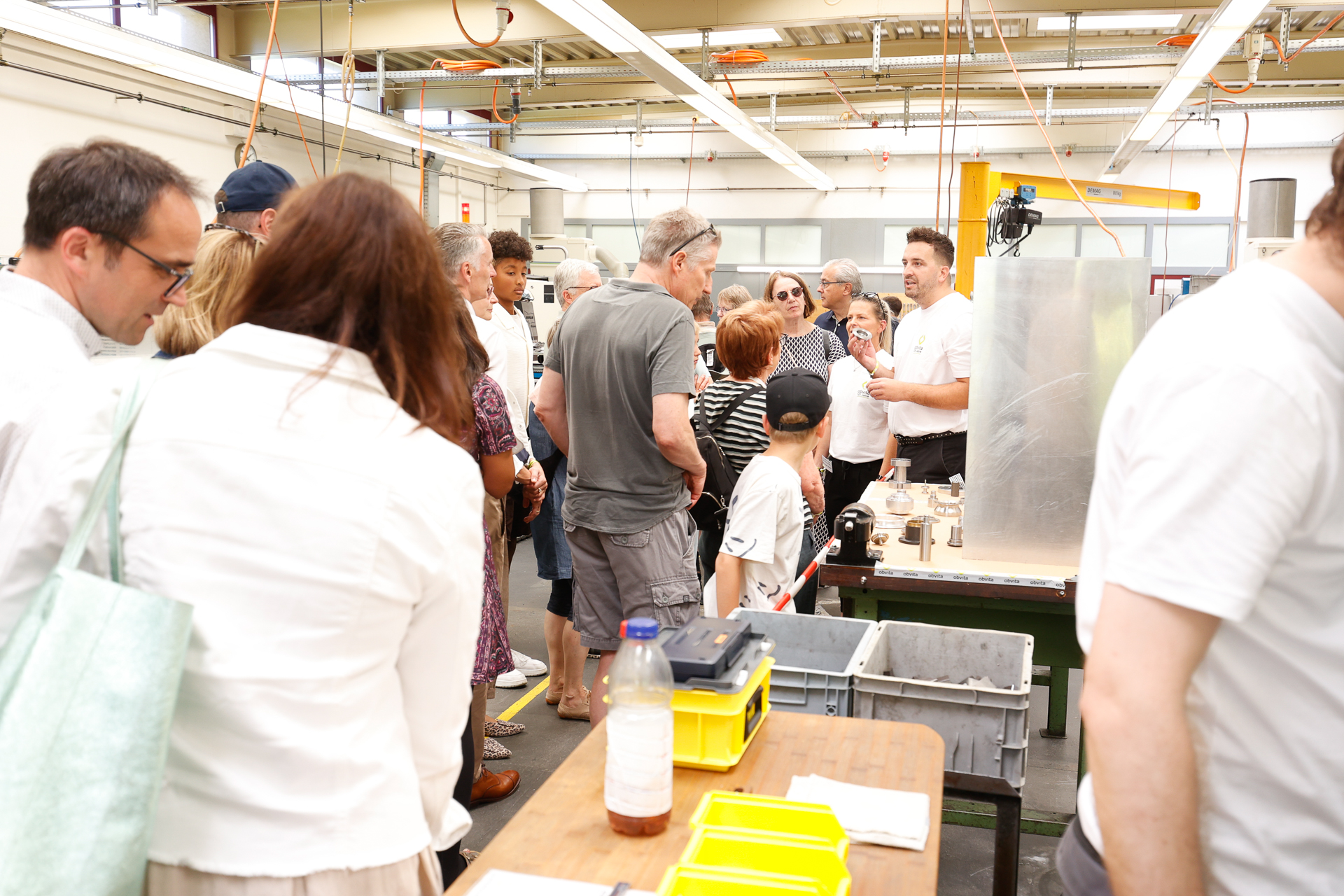Eine Gruppe von Menschen versammelt sich um einen Mann, der in einer Industriehalle eine Vorführung hält. Man sieht Tische mit verschiedenen Werkzeugen und Behältern, und Sonnenlicht durchflutet den geschäftigen Raum.