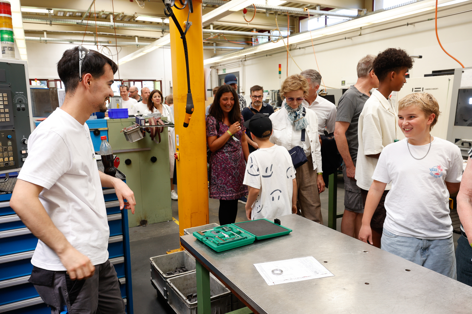 Eine Gruppe von Menschen, darunter Erwachsene und Kinder, versammelt sich in einer Werkstatt, in der verschiedene Maschinen stehen. Einige lächeln und unterhalten sich, und auf der Werkbank im Vordergrund sind Werkzeuge und Papiere zu sehen.