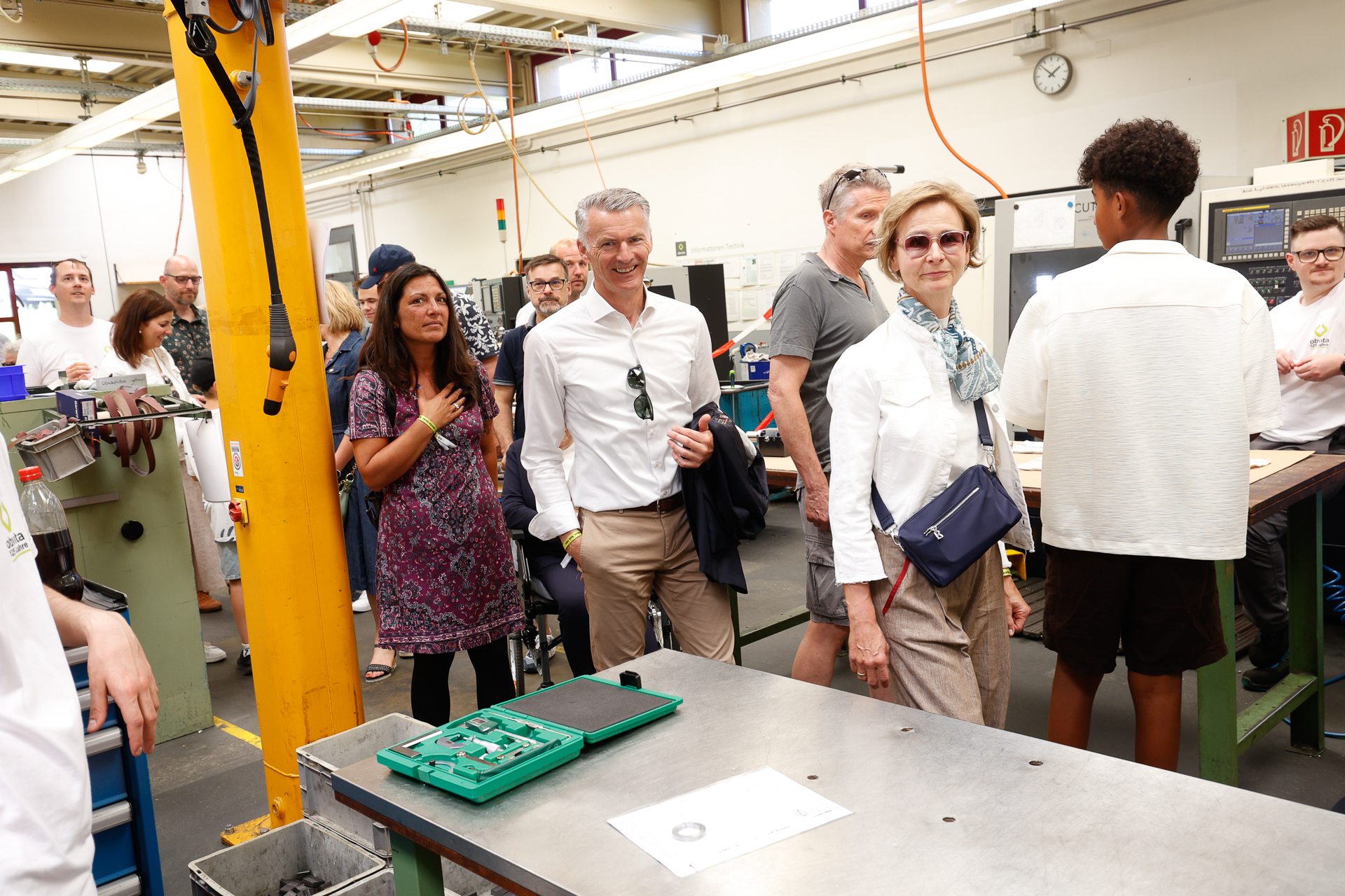 Eine Gruppe von Erwachsenen und Jugendlichen, von denen einige formell gekleidet sind, besichtigt eine Werkstatt mit Maschinen und Werkzeugen; in der hellen, industriellen Umgebung wirken sie interessiert und aufmerksam.