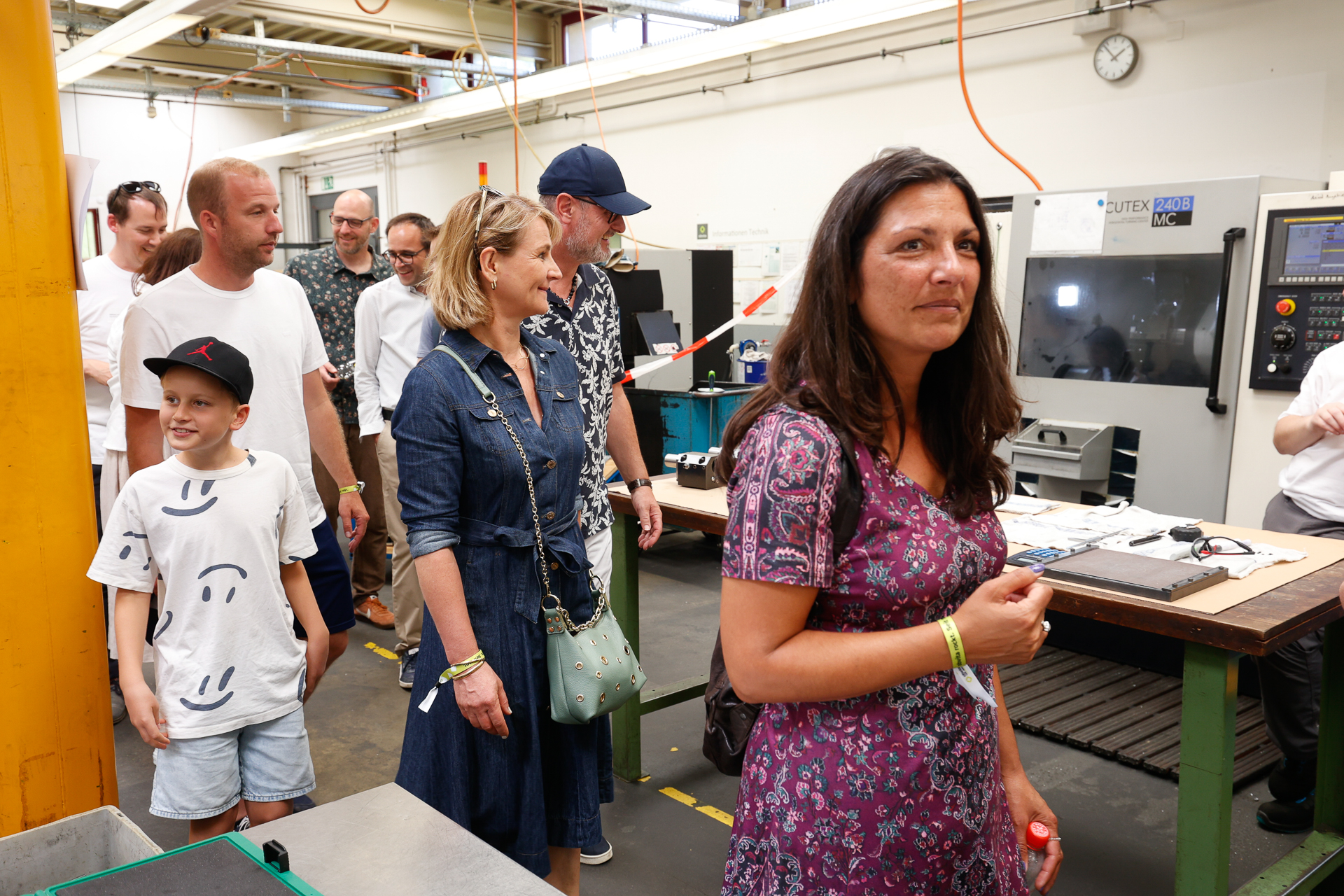 Eine Gruppe von Menschen, darunter Erwachsene und Kinder, geht durch eine hell beleuchtete Industriehalle mit Maschinen und Geräten. Im Vordergrund steht eine Frau in einem violetten Kleid, während sich die anderen im Arbeitsbereich umsehen.
