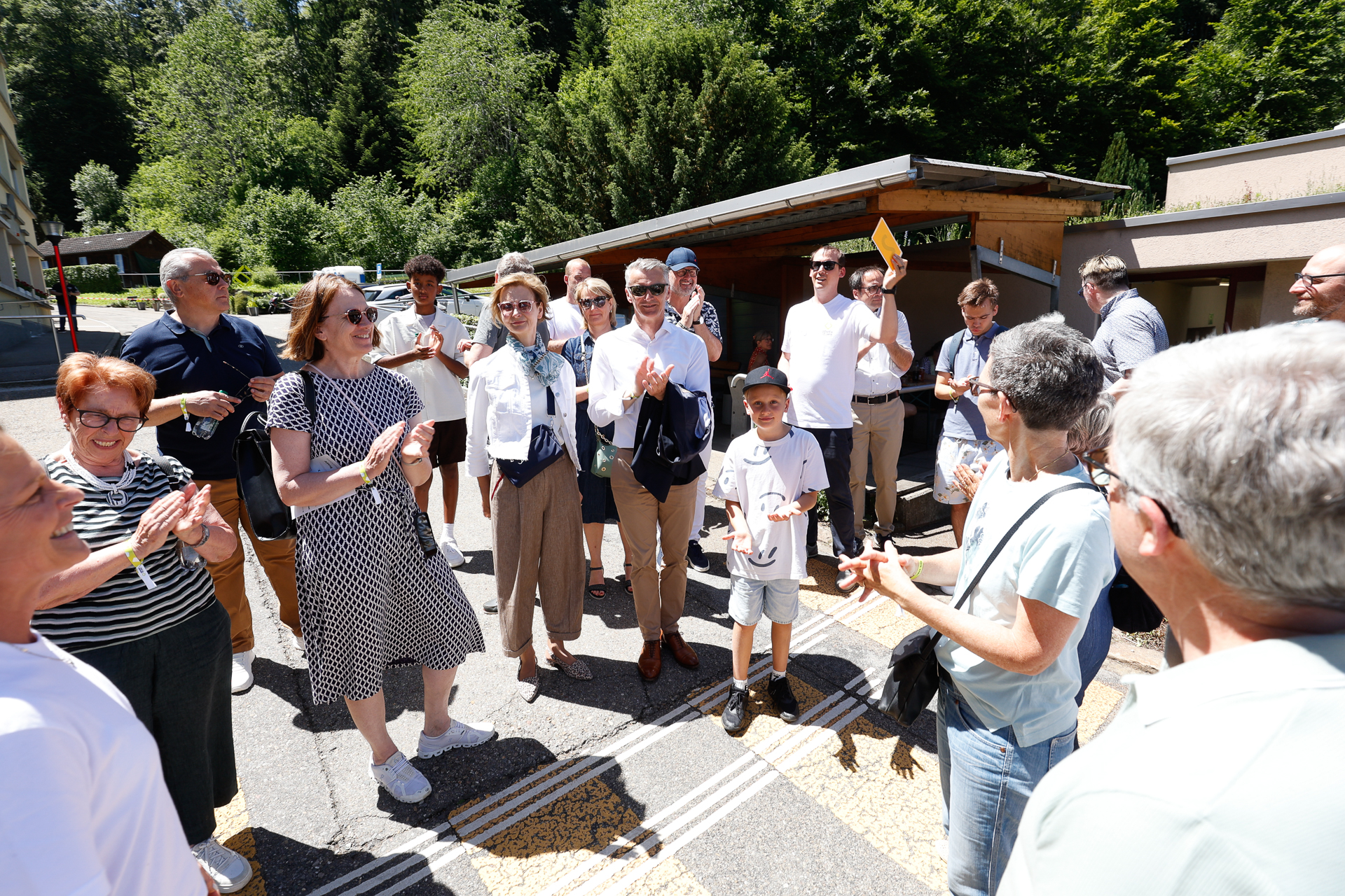 Eine Gruppe von Menschen, darunter Erwachsene und Kinder, steht an einem sonnigen Tag im Freien, lächelt und unterhält sich. Im Hintergrund sind Bäume und Gebäude zu sehen. Einige Personen halten kleine Gegenstände oder Zettel in der Hand.