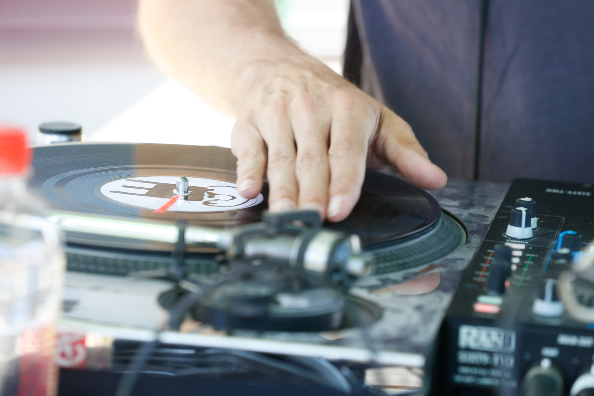 Eine Nahaufnahme der Hand eines DJs, der auf einem Plattenspieler eine Schallplatte „scratcht“, daneben stehen ein Mischpult und weitere Audiogeräte.