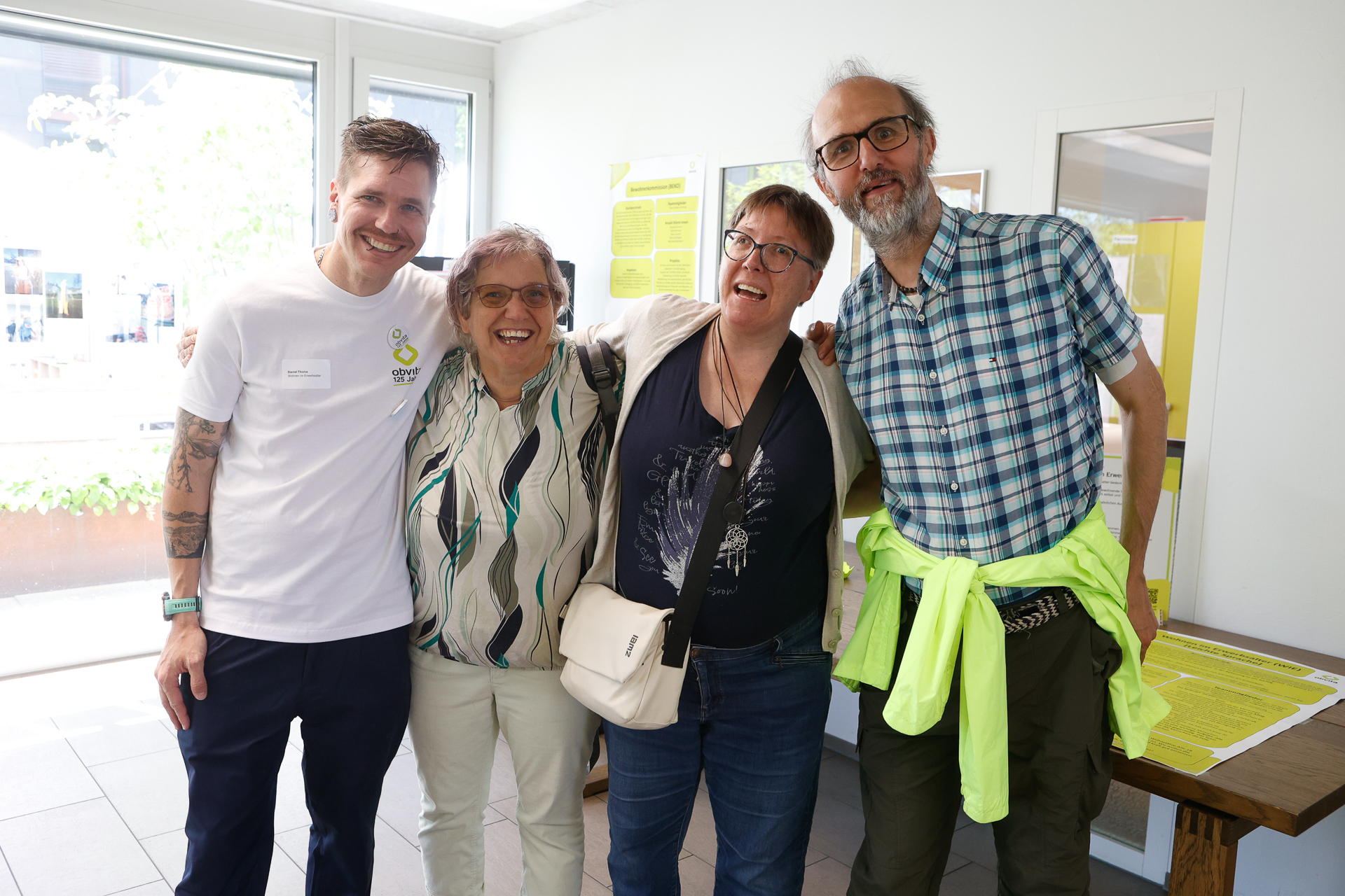 Vier Erwachsene stehen in einem Raum dicht beieinander und lächeln in die Kamera. Ein Mann trägt ein weißes Hemd, die anderen sind leger gekleidet. Eine Person hat eine Tasche dabei, eine andere trägt eine bunte Jacke, die um die Taille gebunden ist. Im Hintergrund sind Informationsplakate zu sehen.