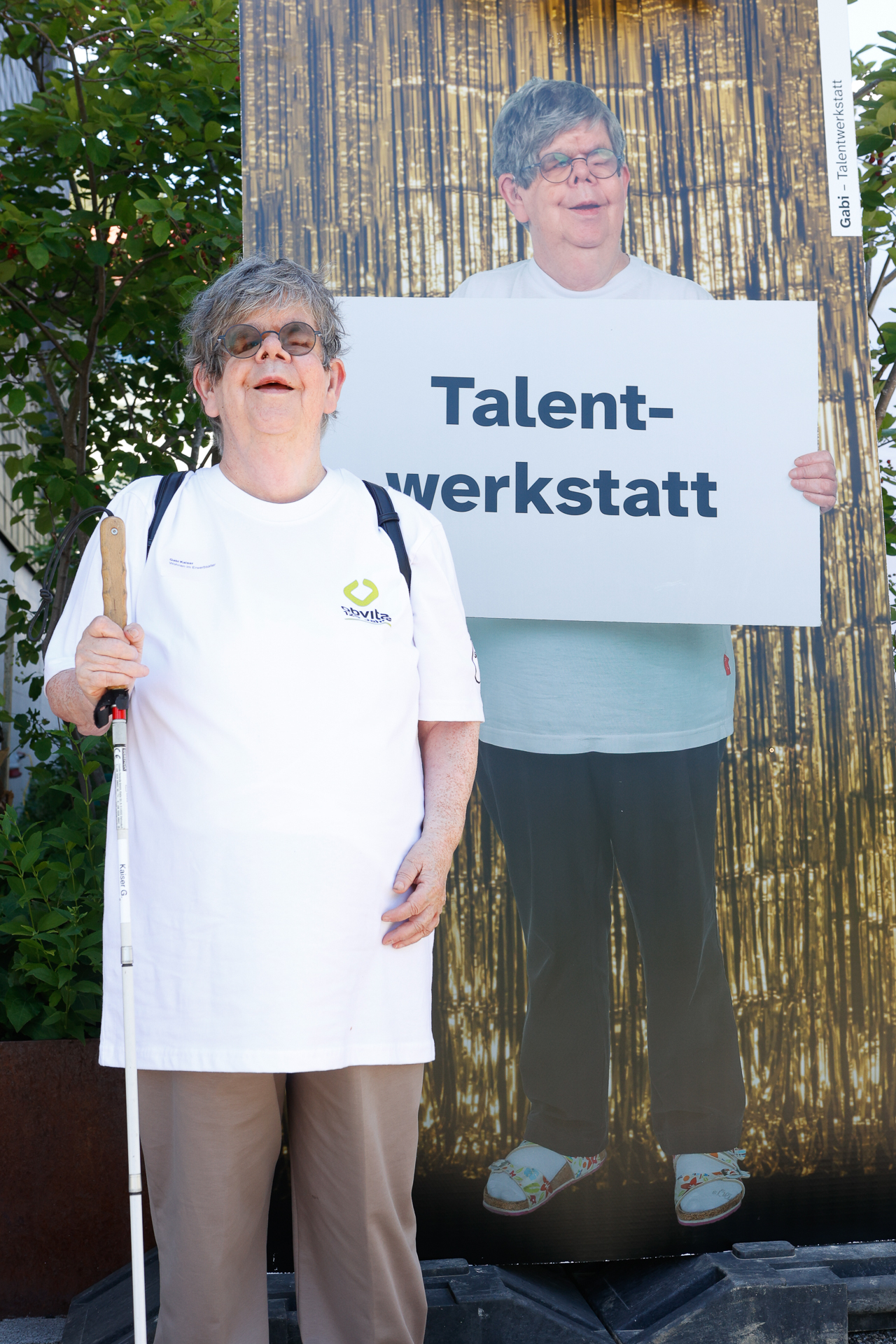 Eine ältere Frau mit grauen Haaren, Brille und einem weißen Blindenstock steht lächelnd neben einer lebensgroßen Pappfigur von sich selbst, die ein Schild mit der Aufschrift „Talentwerkstatt“ in der Hand hält. Sie trägt ein weißes Hemd und eine beige Hose.