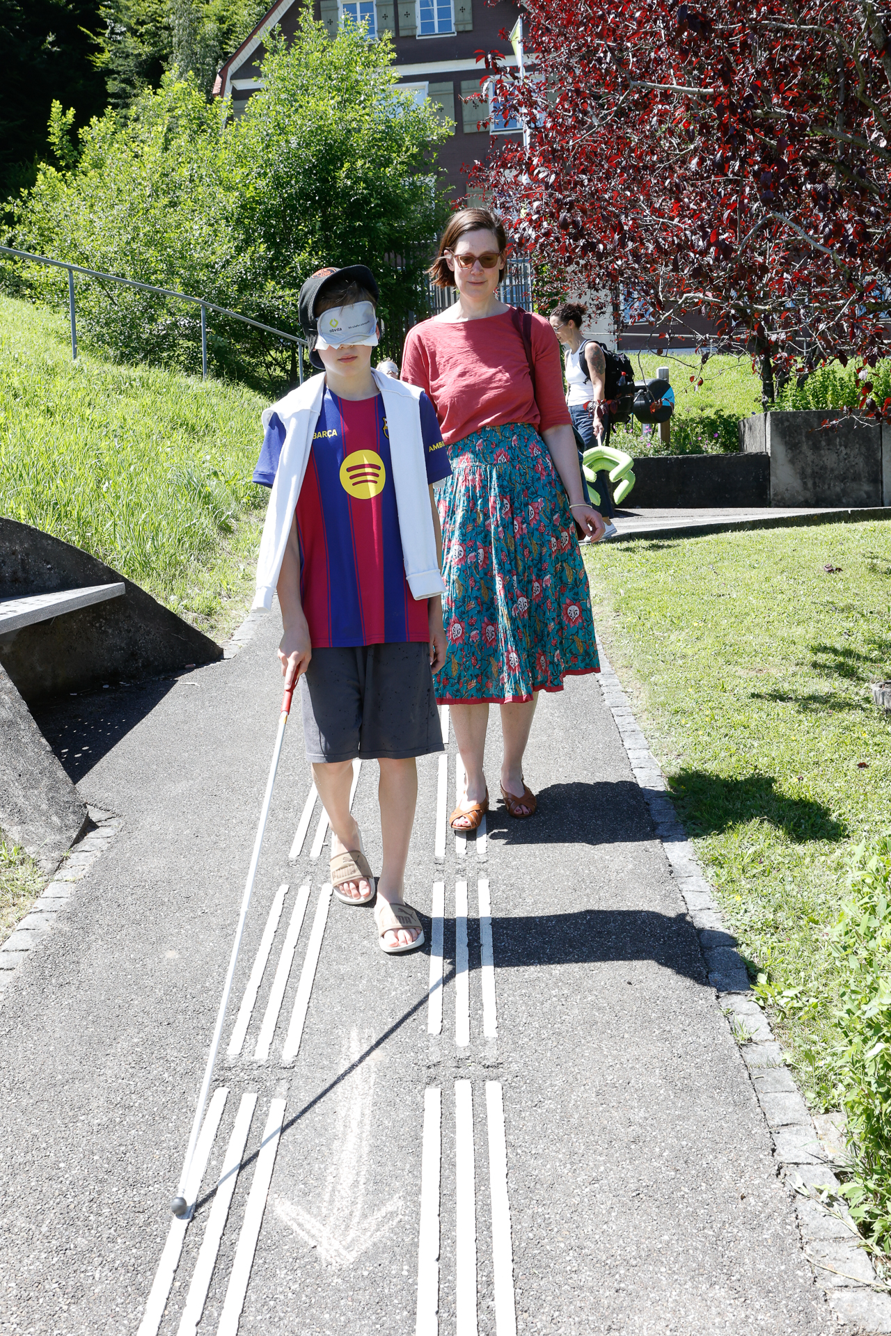 Ein Junge mit einer Augenklappe und einem weißen Blindenstock geht auf einem taktilen Pflasterweg, geführt von einer Frau, die neben ihm hergeht. Sie befinden sich an einem sonnigen Tag im Freien, im Hintergrund sind Gras, Bäume und ein Haus zu sehen.