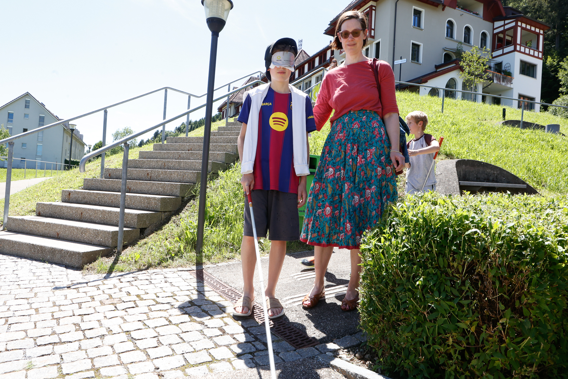 An einem sonnigen Tag steht ein Junge mit einer Augenmaske und einem weißen Blindenstock neben einer Frau an einer Treppe, im Hintergrund ist ein weiterer Junge zu sehen, und um sie herum befinden sich Gebäude und Grünflächen.