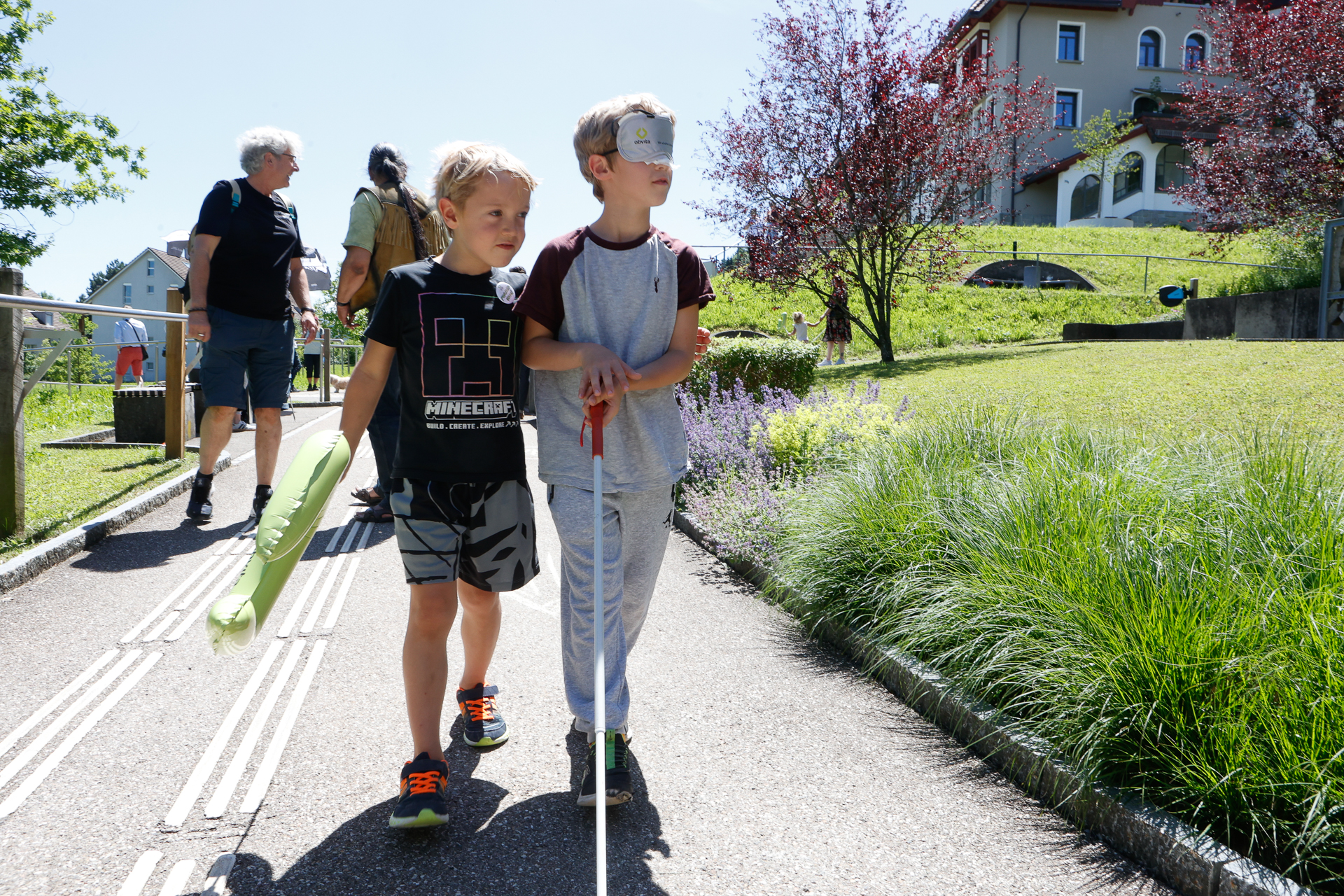 Zwei Jungen gehen auf einem sonnigen Weg; einer trägt eine Augenmaske und benutzt einen weißen Blindenstock, während der andere ihn führt. Im Hintergrund sind in der Nähe eines Gebäudes Erwachsene und Grünflächen zu sehen.