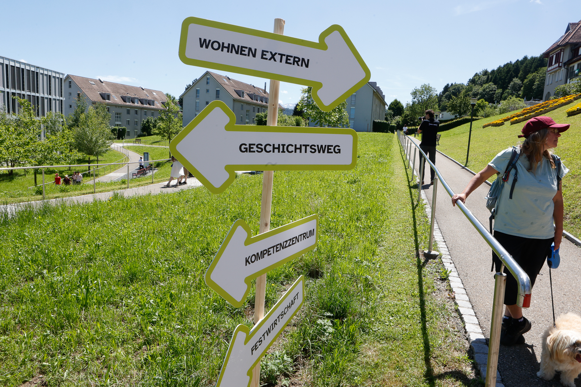 Eine Frau geht mit einem Hund auf einem sonnigen Weg in der Nähe eines Wegweisers spazieren, auf dem vier Pfeile in deutscher Sprache zu verschiedenen Orten zeigen. Im Hintergrund sind Wohnhäuser, grünes Gras und Bäume zu sehen.