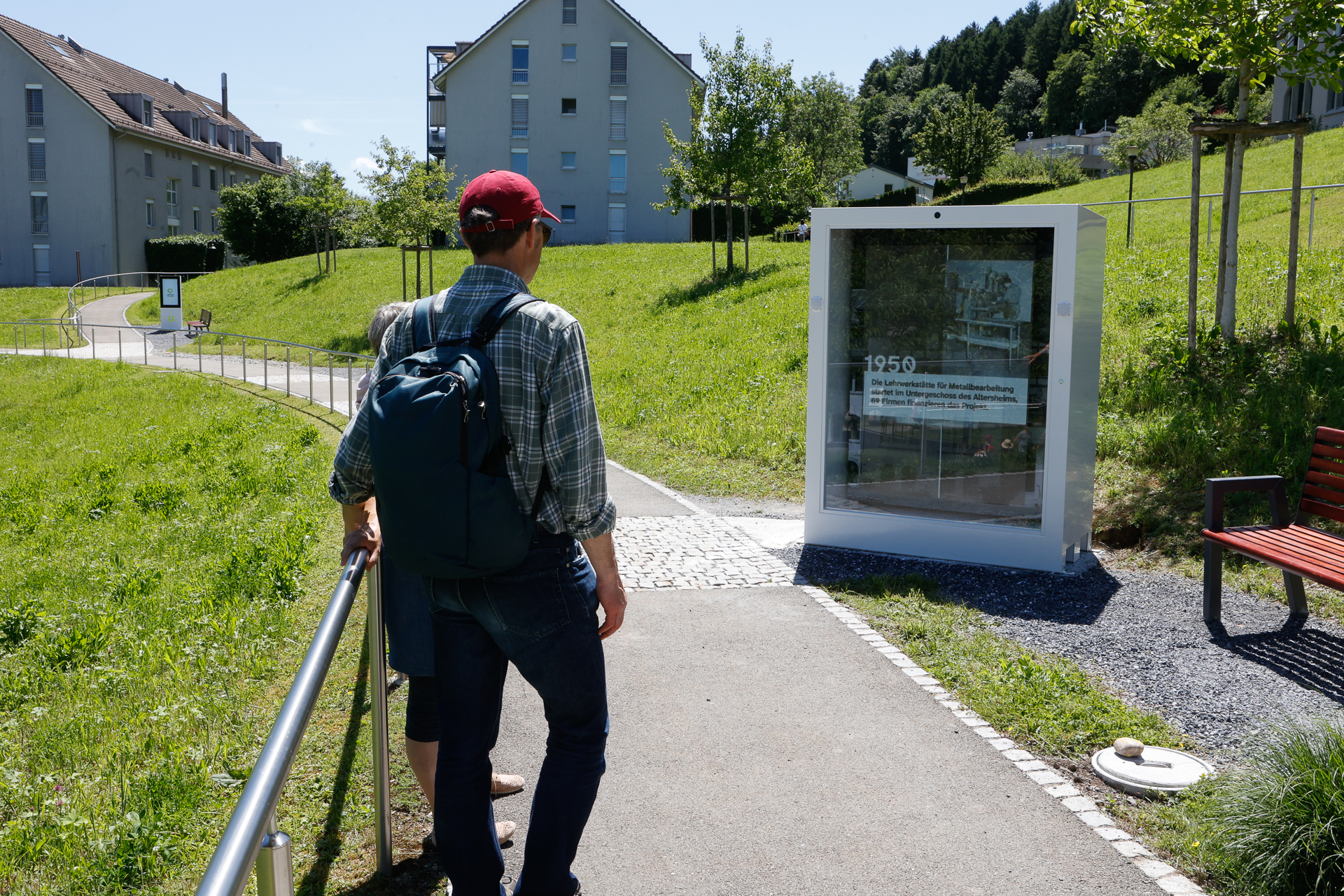 Eine Person mit roter Mütze und Rucksack steht auf einem gepflasterten Weg und liest eine Informationstafel im Freien, auf der „1950“ steht. Der Weg ist von Rasenflächen und nahegelegenen Wohngebäuden gesäumt.