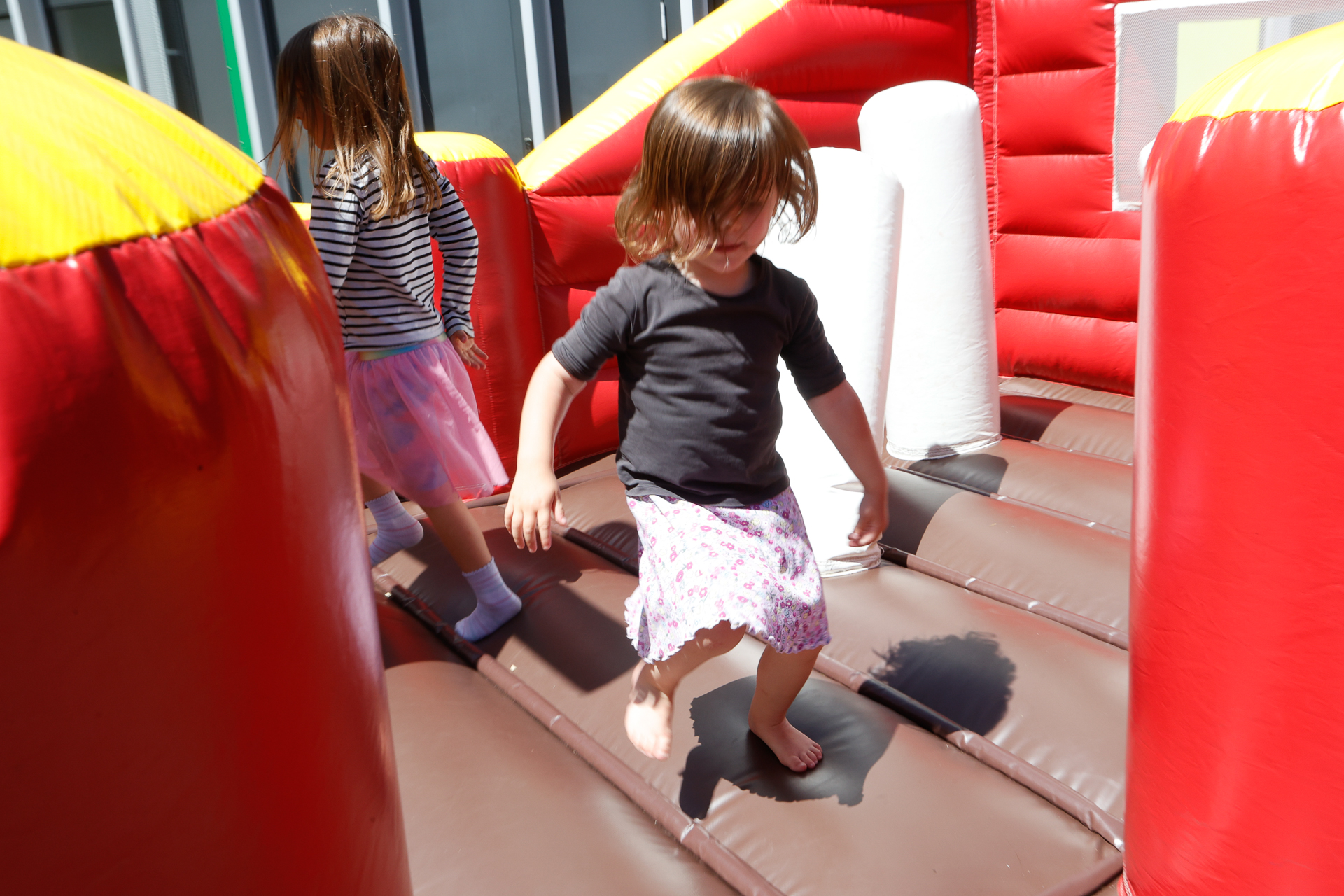 An einem sonnigen Tag spielen zwei kleine Kinder in einer rot-braunen aufblasbaren Hüpfburg. Ein Kind schaut nach vorne, das andere hat den Rücken zu; beide tragen Röcke und haben viel Spaß dabei.