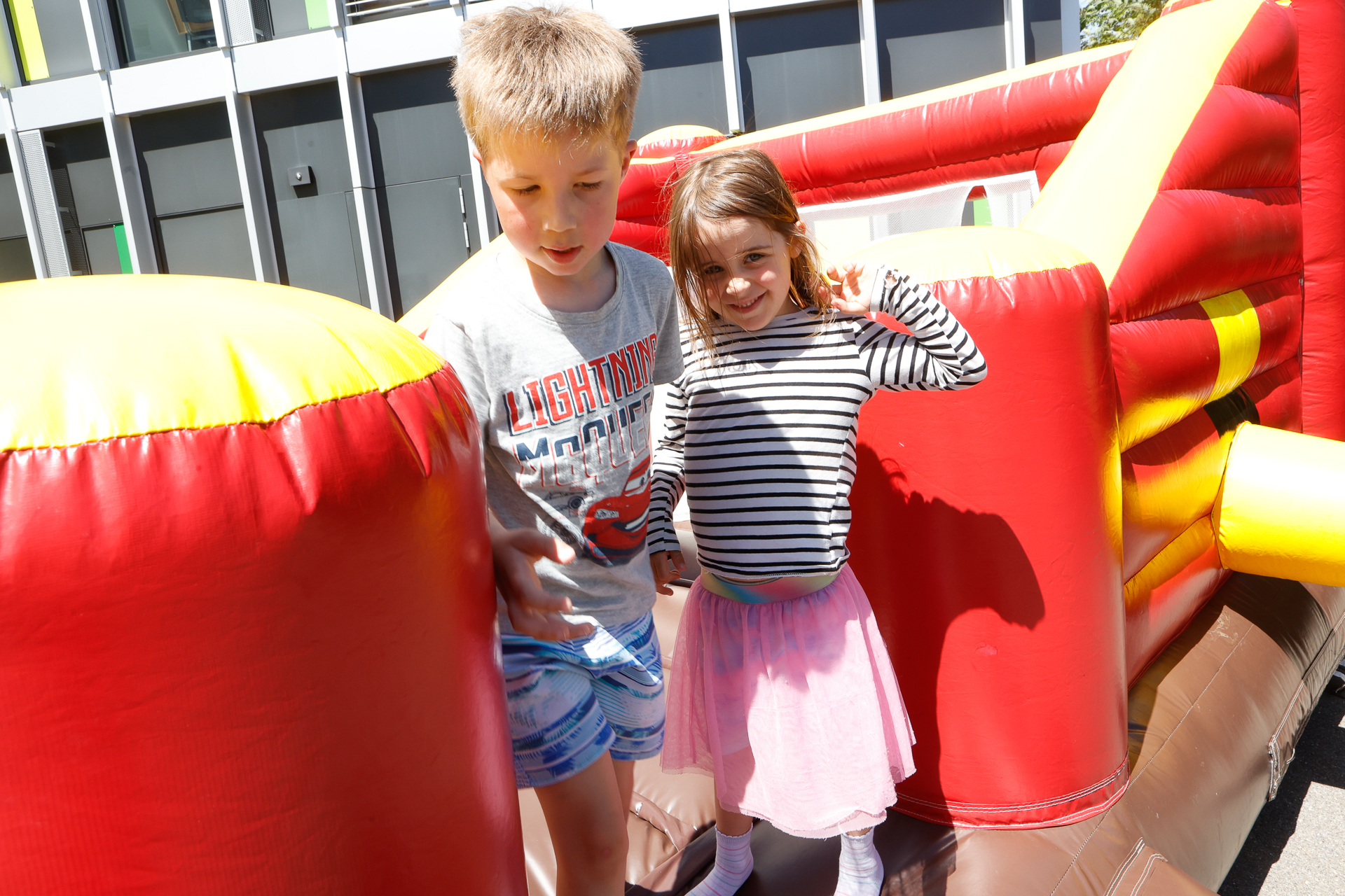 An einem sonnigen Tag stehen zwei Kinder auf einem rot-gelben aufblasbaren Hindernisparcours im Freien und lächeln. Der Junge trägt ein graues T-Shirt und blaue Shorts; das Mädchen trägt ein gestreiftes Oberteil und einen rosa Rock.