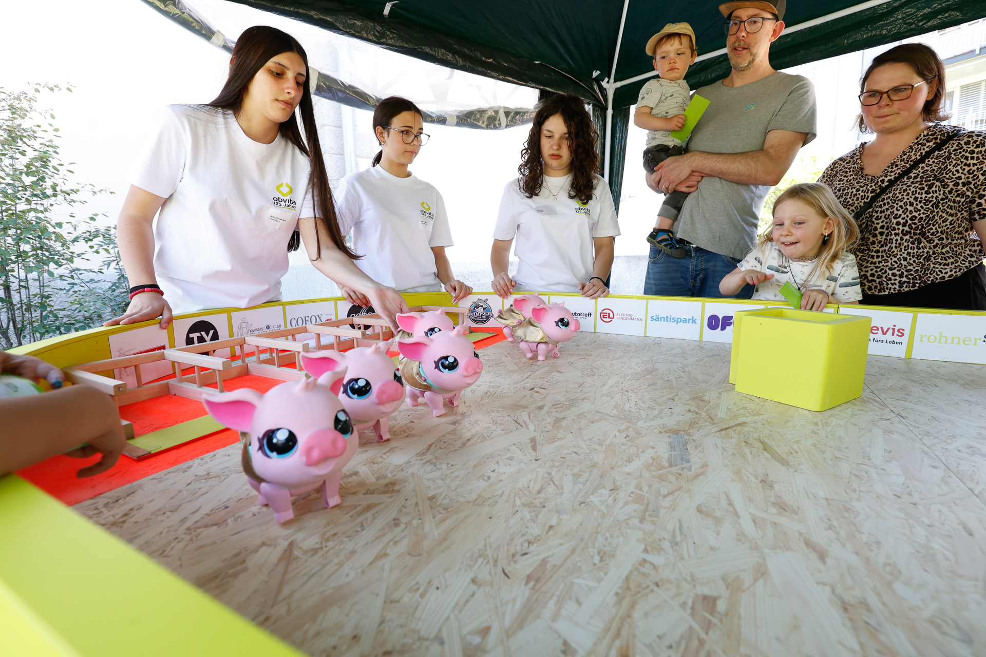 Eine Gruppe von Kindern und Erwachsenen versammelt sich um ein Brettspiel, bei dem mehrere Spielzeugschweine auf einer Holzplatte stehen; einige Teilnehmer tragen weiße Hemden, andere schauen zu und lächeln.