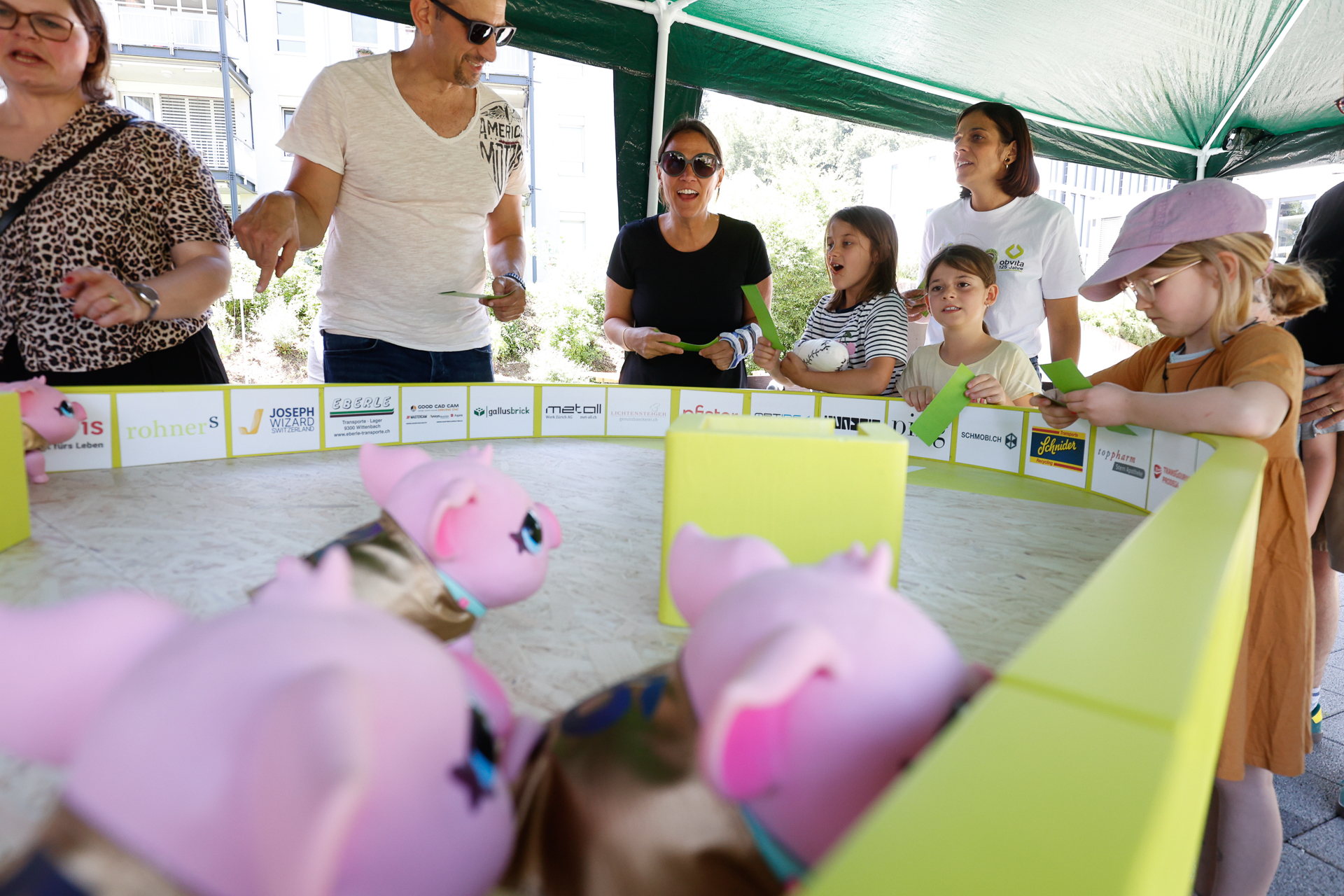 Eine Gruppe von Erwachsenen und Kindern steht um eine kleine, eingezäunte Arena herum und beobachtet, wie rosa Spielzeugschweine auf sandigem Untergrund um die Wette laufen; sie wirken begeistert und fasziniert. Die Szene spielt sich unter einem grünen Zelt im Freien ab.