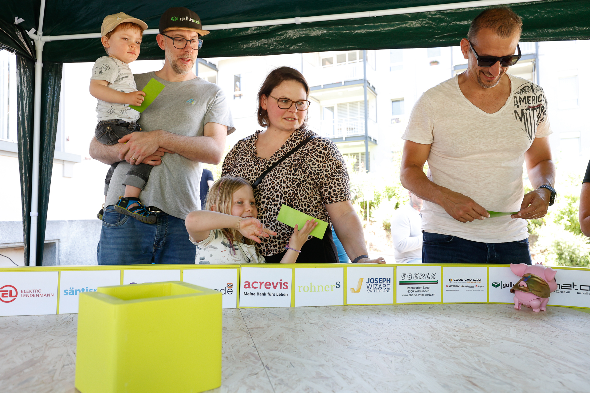 Vier Erwachsene und zwei Kinder stehen unter einem Vordach, halten grüne Karten in der Hand und blicken auf eine gelbe Schachtel auf einem Tisch. An der Seite des Tisches sind Sponsorenlogos zu sehen, und eines der Kinder streckt die Hand nach der Schachtel aus.