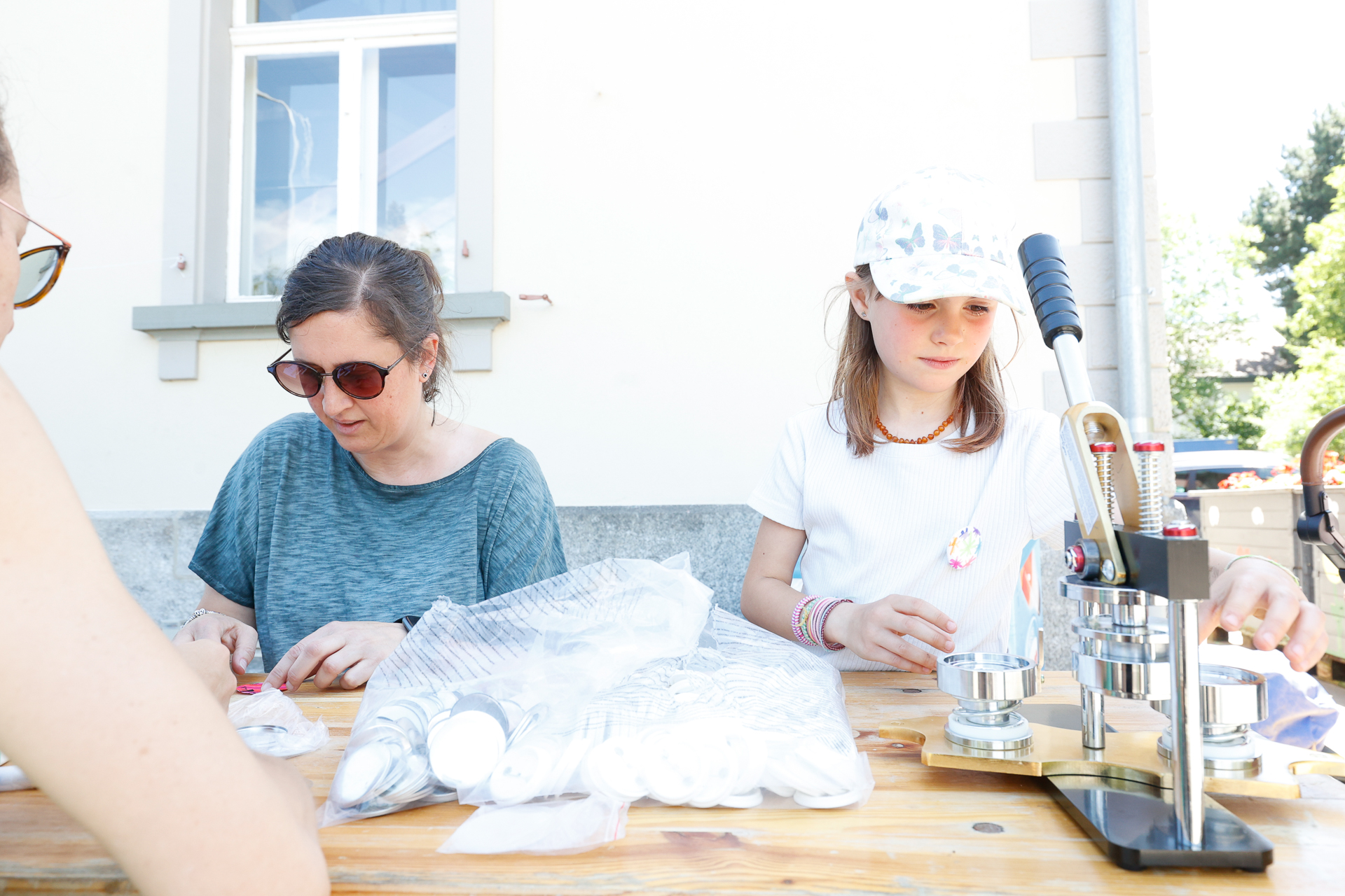 Zwei Personen sitzen an einem Tisch im Freien und stellen mit einer Knopfmaschine Ansteckknöpfe her. Die eine trägt eine Sonnenbrille und ein blaues Hemd, die andere ein weißes Hemd und eine Mütze. Auf dem Tisch liegen Knopfteile und Zubehör ausgebreitet.