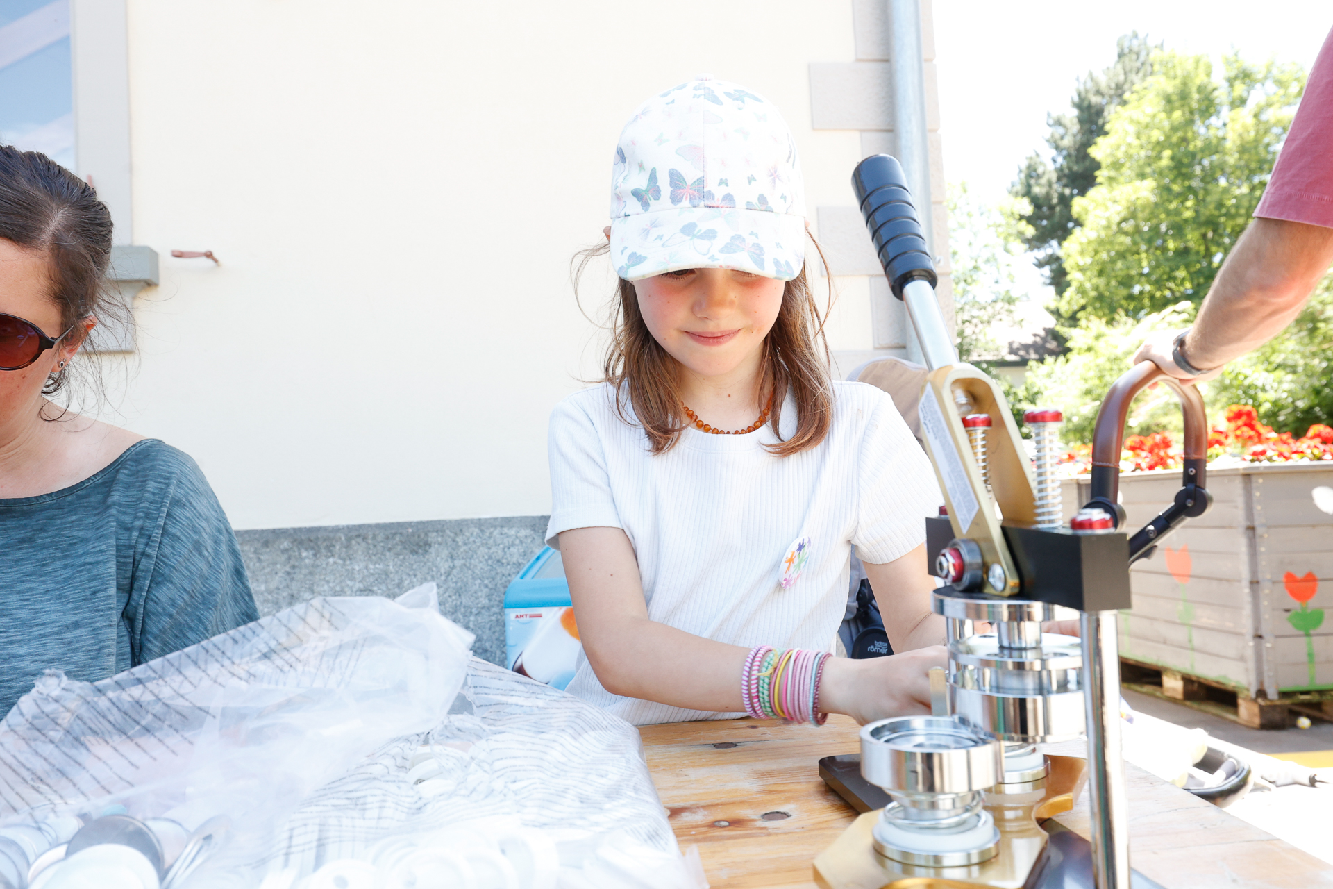 Ein junges Mädchen mit weißer Mütze und T-Shirt sitzt draußen an einem Holztisch und bedient eine Knopfmaschine. Sie ist ganz in ihre Arbeit vertieft, während auf dem Tisch Knopfteile und Zubehör verstreut liegen.