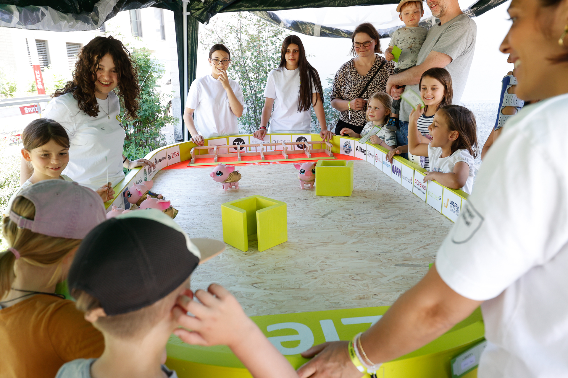 Eine Gruppe von Kindern und Erwachsenen versammelt sich bei einer Veranstaltung im Freien unter einem Zelt um ein Brettspiel, bei dem kleine rosa Schweinchenroboter auf einer Holzplatte mit Hindernissen zu sehen sind. Alle wirken begeistert und glücklich.