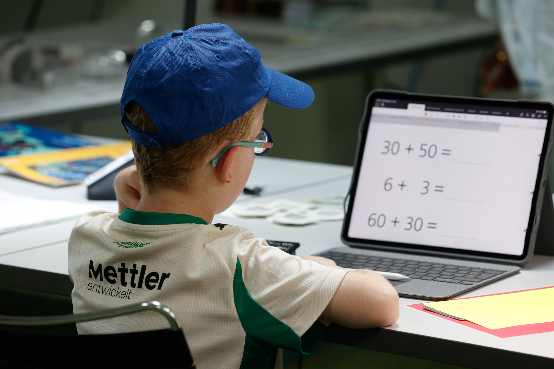 Ein kleiner Junge mit einer blauen Mütze und einer Brille sitzt an einem Schreibtisch und schaut auf ein Tablet, auf dem einfache Rechenaufgaben angezeigt werden. Er trägt ein Sportshirt, auf dessen Rücken „Mettler entwickelt“ steht. Auf dem Schreibtisch liegen Schulsachen.