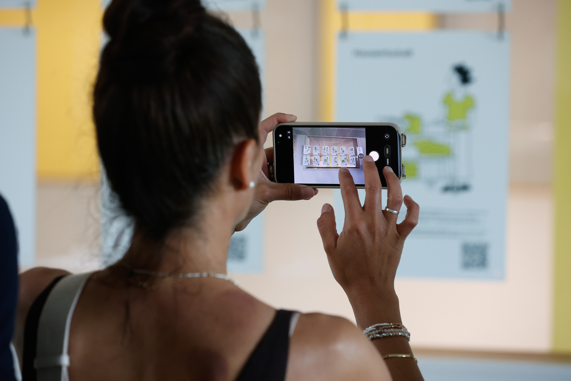 Eine Frau mit dunklem Haar, das zu einem Dutt zusammengebunden ist, fotografiert mit ihrem Smartphone Plakate, die an einer Wand hängen. Die Plakate zeigen Illustrationen und Text, während auf dem Bildschirm ihres Smartphones eine Nahaufnahme eines der Plakate zu sehen ist.