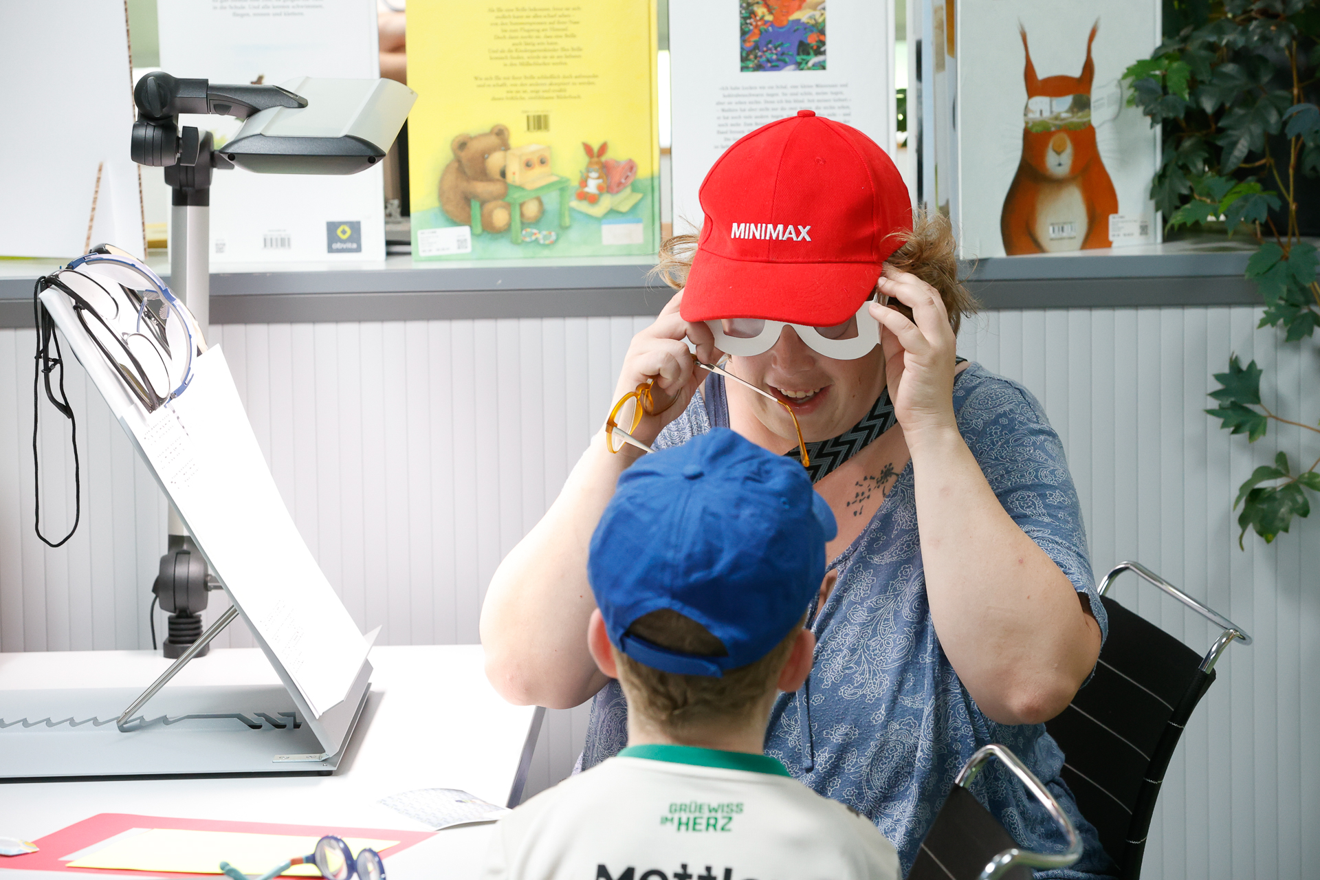 Eine Frau mit einer roten Mütze und einer großen Spielzeugbrille lächelt einen kleinen Jungen mit einer blauen Mütze an, der ihr an einem Tisch in einem bunten, kinderfreundlichen Raum gegenüber sitzt.