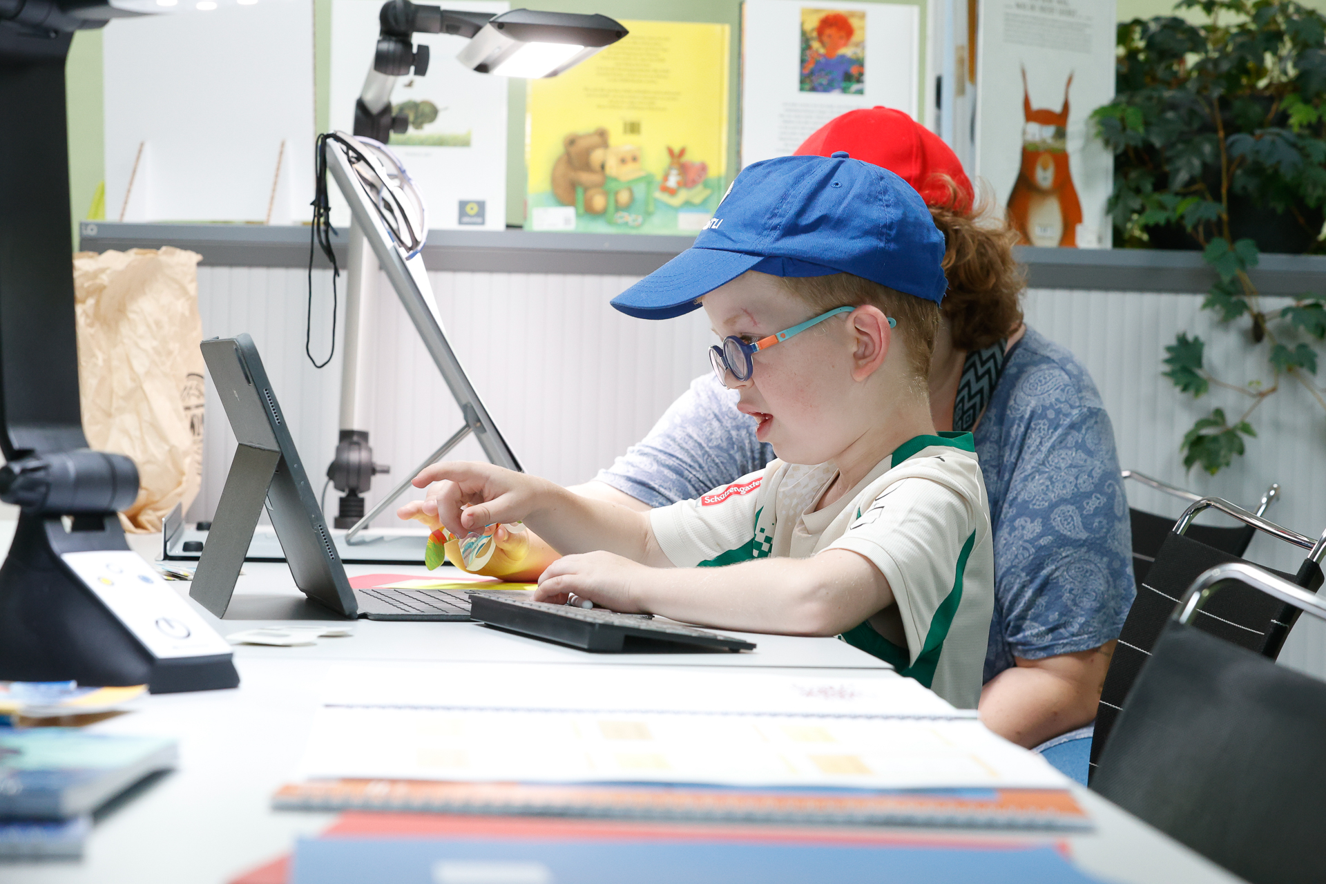 Ein kleiner Junge mit blauer Mütze und Brille bedient ein Tablet mit Hilfe eines Erwachsenen, der neben ihm in einem hell erleuchteten Raum sitzt, umgeben von Büchern, Papieren und Lernmaterialien.
