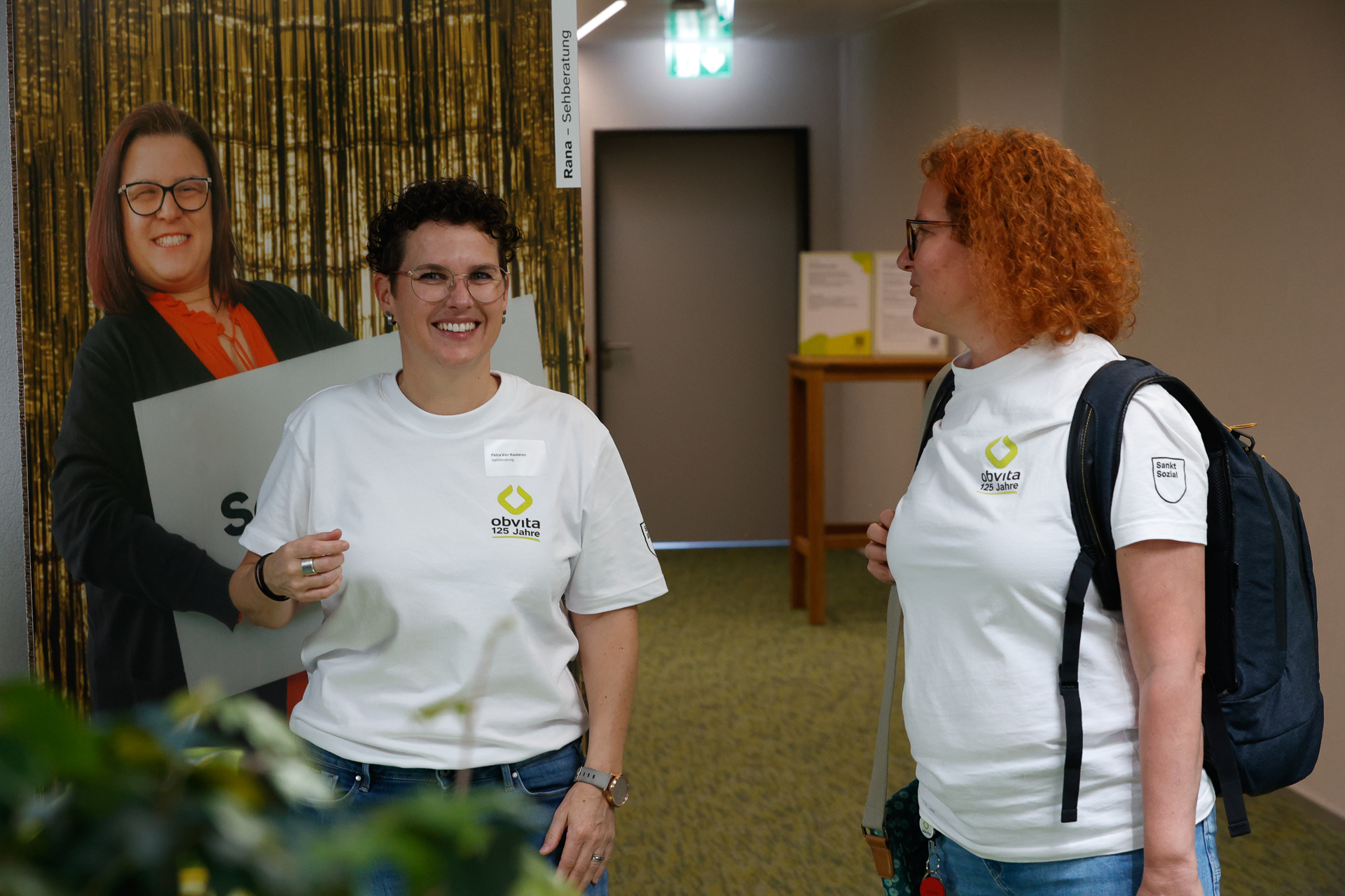 Zwei Frauen in weißen Shirts mit „olyata“-Logos unterhalten sich und lächeln in einem Raum, während sie neben einer lebensgroßen Pappfigur einer Frau stehen. Eine der Frauen trägt eine Brille und einen Rucksack; im Hintergrund sind ein Flur und eine Tür zu sehen.