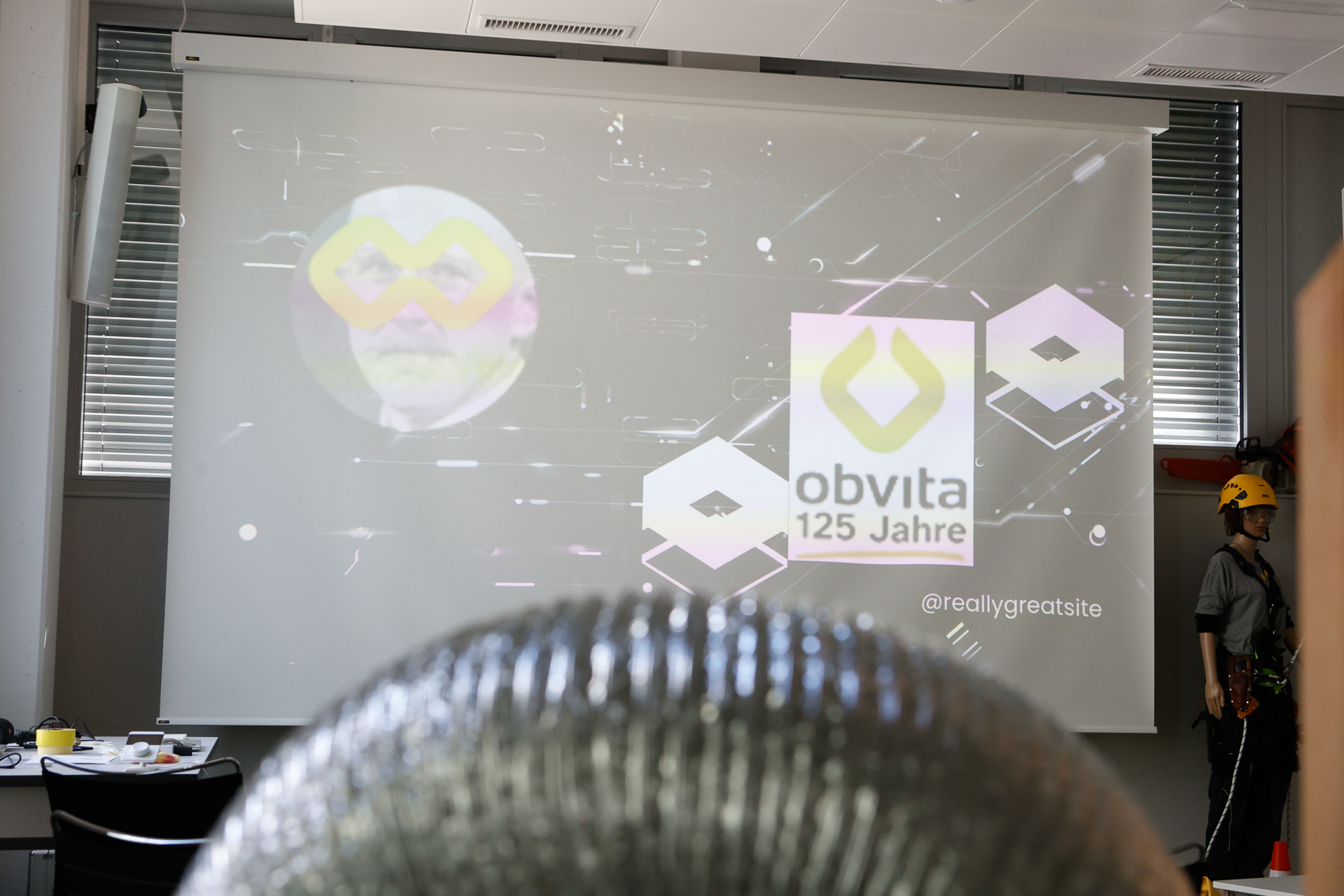 Auf einer Präsentationsfolie steht „obvita 125 Jahre“ neben einem gelben Logo. Rechts steht eine Person mit Helm, und im Vordergrund ist die glänzende Oberseite eines Gegenstands zu sehen. Links stehen Tische mit Gegenständen darauf.