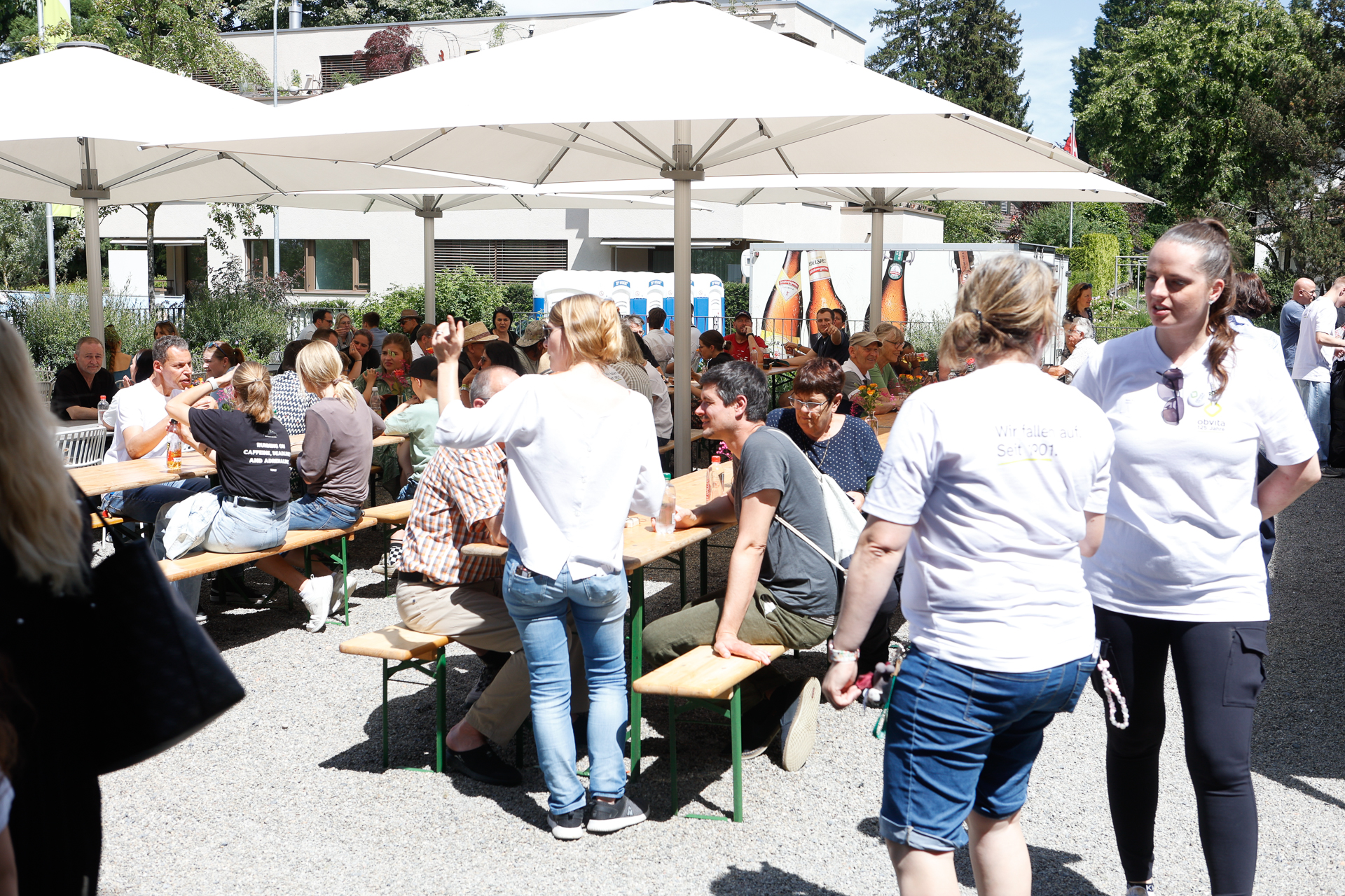 Die Menschen haben sich draußen unter großen weißen Sonnenschirmen versammelt, sitzen an Holztischen und auf Bänken, unterhalten sich und genießen den sonnigen Tag. Im Vordergrund stehen zwei Frauen in weißen Blusen, während sich im Hintergrund andere unterhalten.