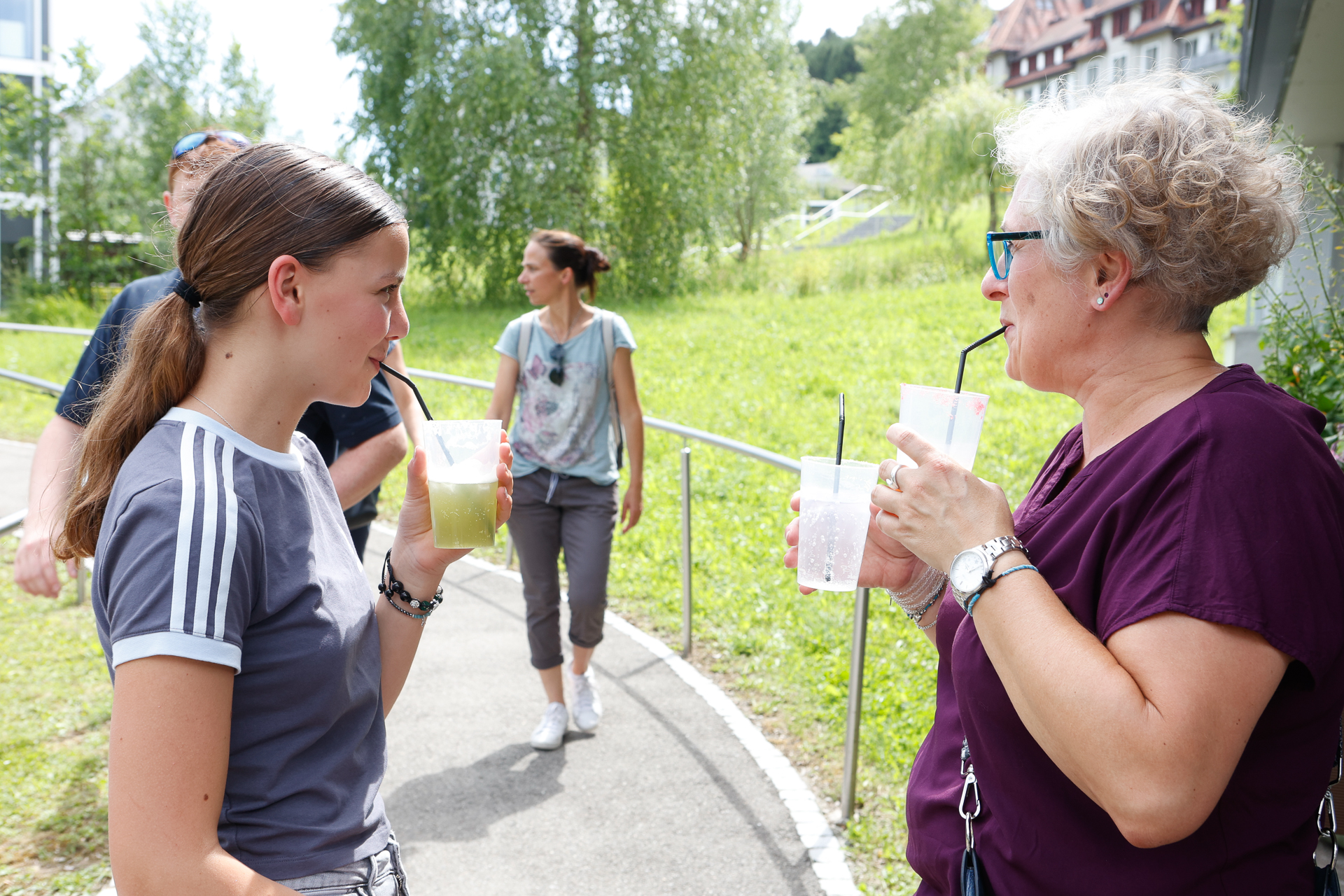 Zwei Frauen stehen an einem sonnigen Tag draußen, trinken etwas und unterhalten sich. Im Hintergrund sind Menschen zu sehen, die auf einem Weg spazieren gehen, umgeben von grünem Gras und Bäumen. Die Atmosphäre wirkt entspannt und gesellig.