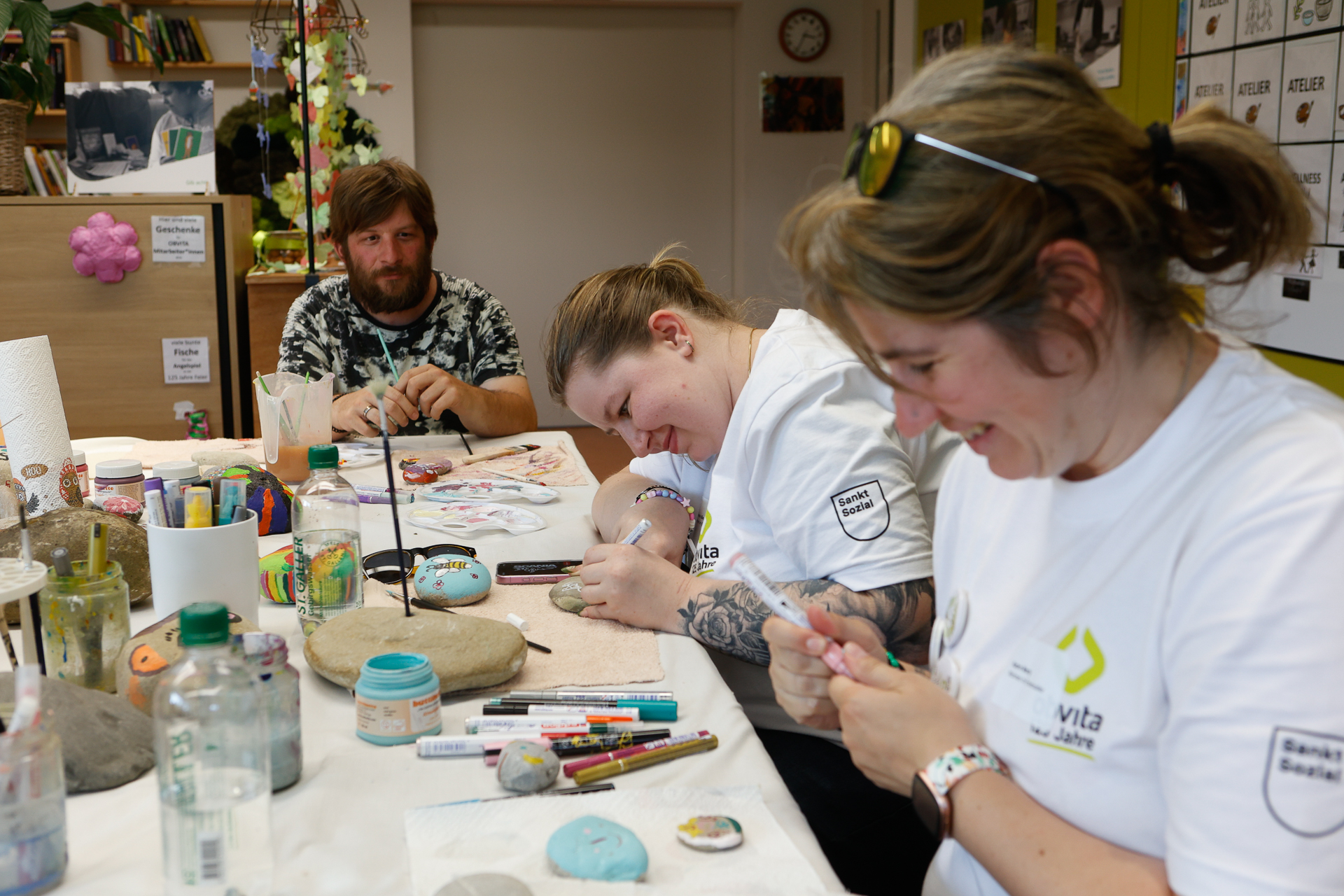 Drei Menschen sitzen an einem Tisch, der mit Malutensilien bedeckt ist, lächeln und bemalen Steine. Zwei Frauen in weißen Hemden konzentrieren sich auf ihre Kunstwerke, während ein Mann im Hintergrund zusieht und lächelt. Die Szene spielt sich in einem hellen und kreativen Raum ab.