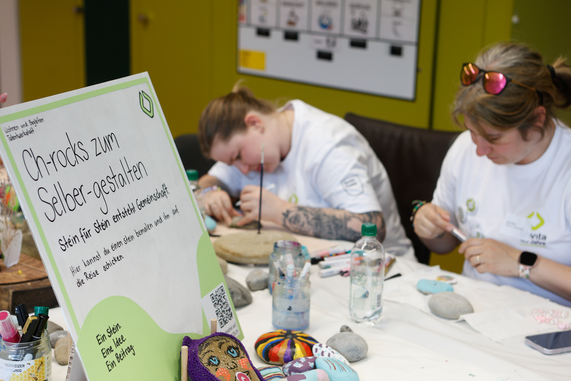 Zwei Personen sitzen an einem Tisch und bemalen Steine, umgeben von Malutensilien. Ein großes Schild auf Deutsch im Vordergrund lädt die Teilnehmer dazu ein, ihren eigenen Stein zu verzieren. Auf dem Tisch sind bemalte Steine und Filzstifte zu sehen.