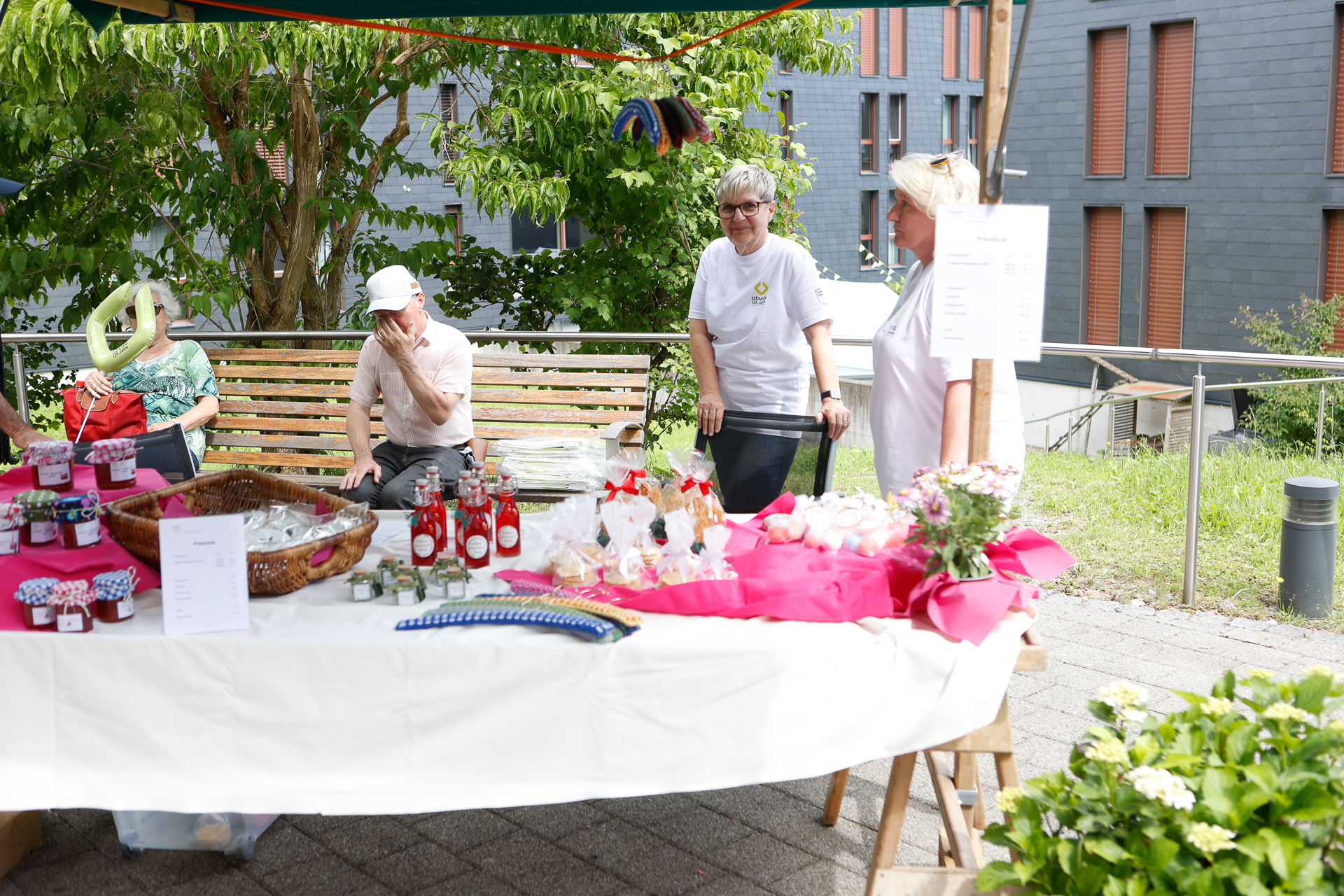 Ein Marktstand im Freien, hinter dessen Tisch zwei Frauen stehen und selbstgemachte Waren wie Gläser, Gebäck und Blumen ausstellen; ein Mann sitzt auf einer Bank in der Nähe und verdeckt sein Gesicht teilweise mit der Hand.