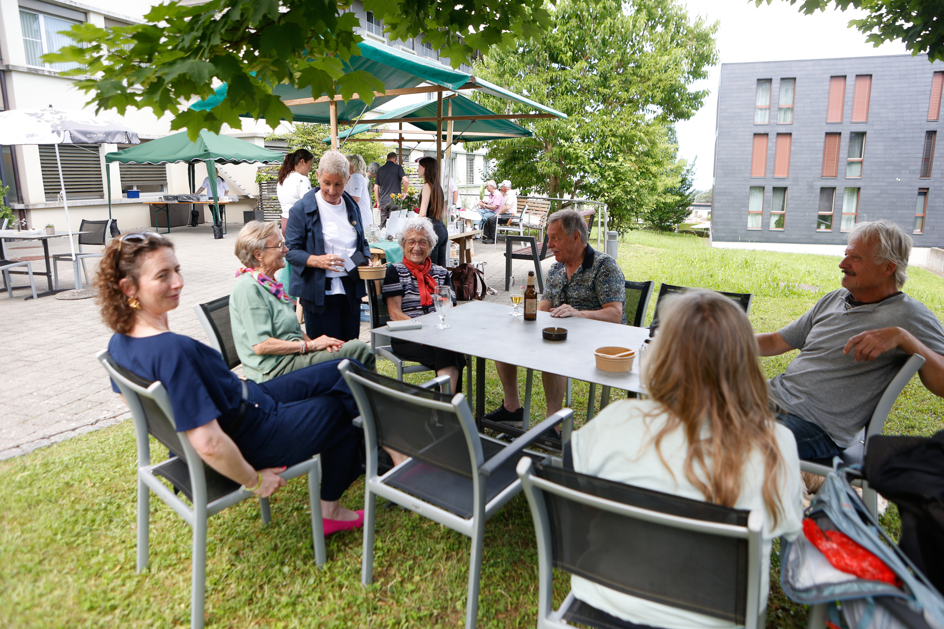 Eine Gruppe älterer Menschen sitzt im Freien unter Bäumen und Sonnenschirmen auf einer Rasenfläche um einen Tisch herum und unterhält sich, während sie ein ungezwungenes Beisammensein in der Nähe moderner Gebäude genießt.