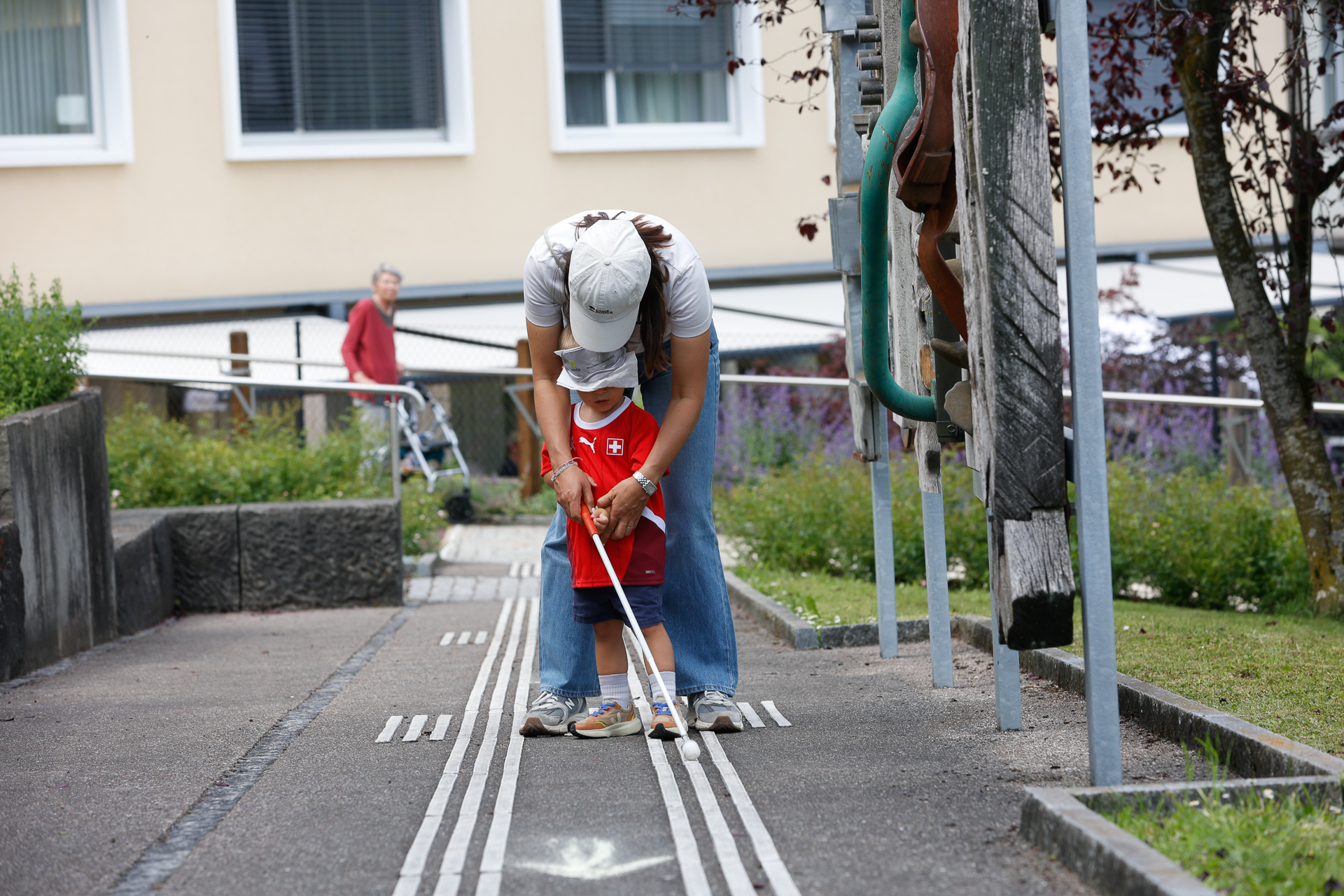 Ein Erwachsener hilft einem kleinen Kind, das eine Augenbinde trägt und einen weißen Blindenstock in der Hand hält, dabei, dem taktilen Bodenbelag auf einem Weg im Freien zu folgen, und veranschaulicht so die Orientierungsfähigkeiten von Menschen mit Sehbehinderungen.