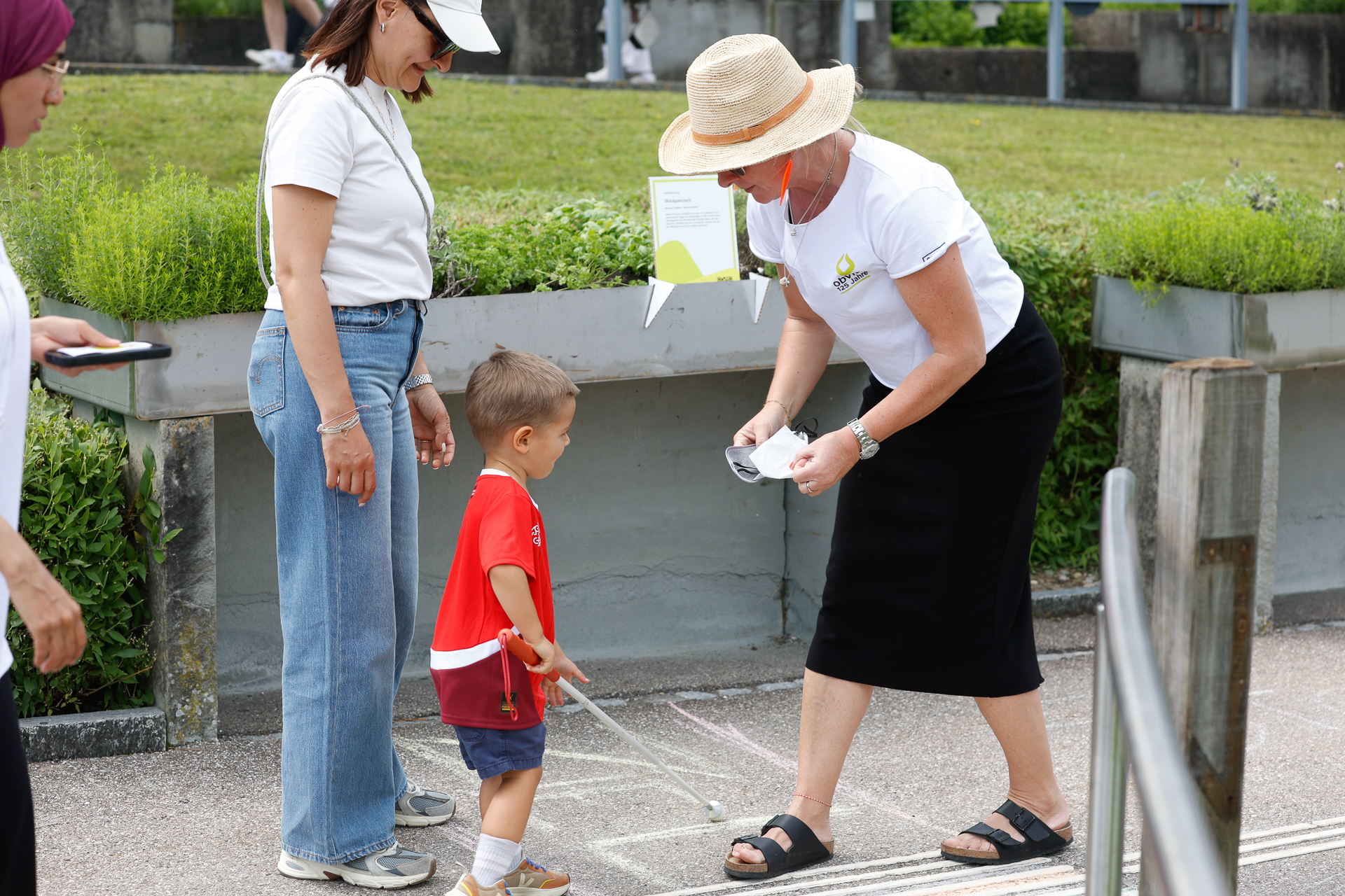Eine Frau mit Hut hilft einem kleinen Jungen, der einen weißen Blindenstock in der Hand hält, mit Kreide auf den Boden zu malen, während eine andere Frau in der Nähe steht – draußen an einem sonnigen Tag.
