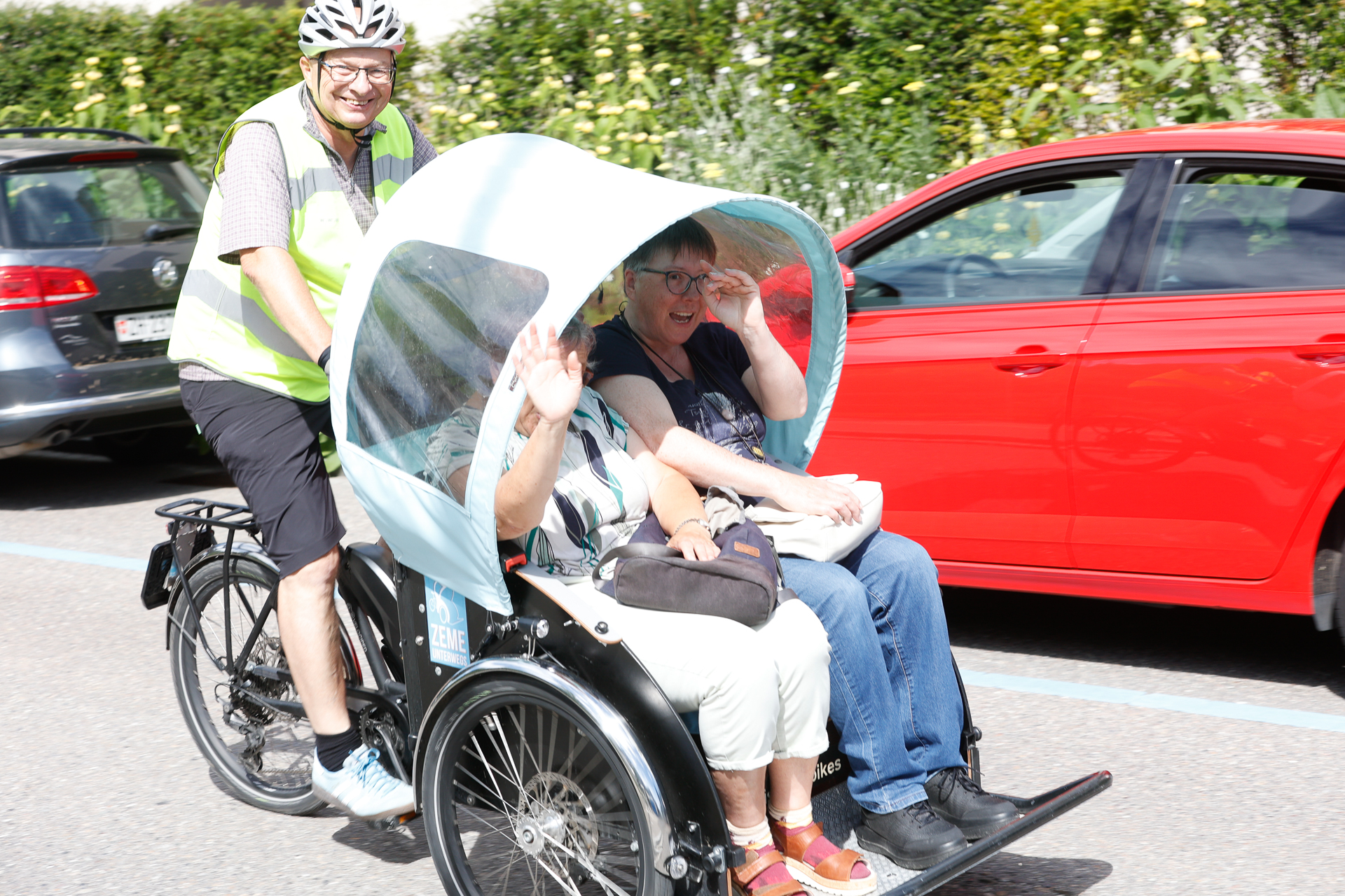 Ein Mann mit Helm und Warnweste tritt in die Pedale einer Dreirad-Rikscha, auf der zwei lächelnde Frauen sitzen – eine von ihnen winkt –, während im Hintergrund eine Straße mit geparkten Autos zu sehen ist.