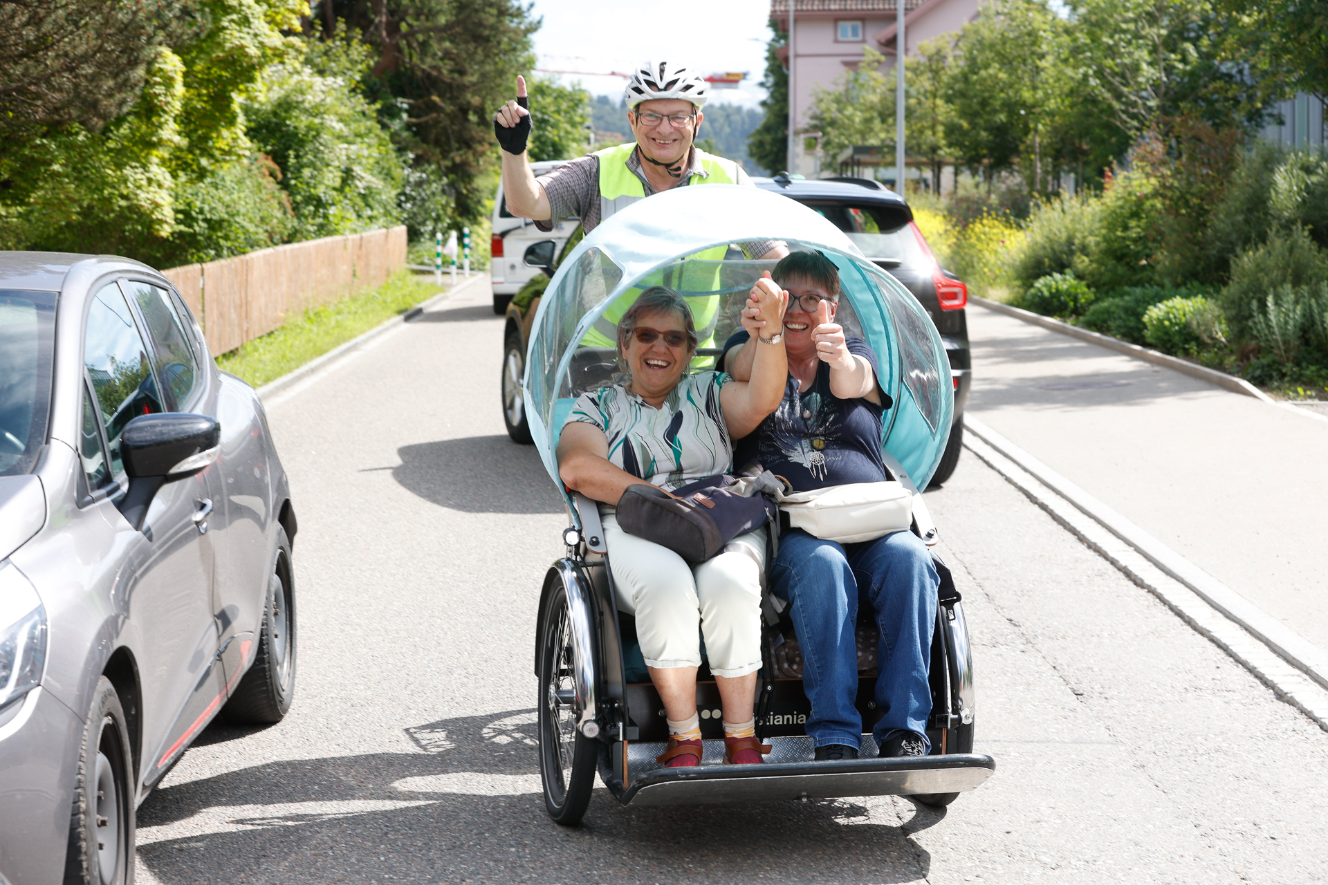 Zwei ältere Frauen sitzen in einer überdachten Rikscha auf einer sonnigen Straße, lächeln und zeigen den Daumen nach oben, während ein Mann mit Helm und Warnweste hinter ihnen herradelt, ebenfalls lächelnd und den Daumen nach oben zeigend. Autos und Grünflächen säumen die Straße.