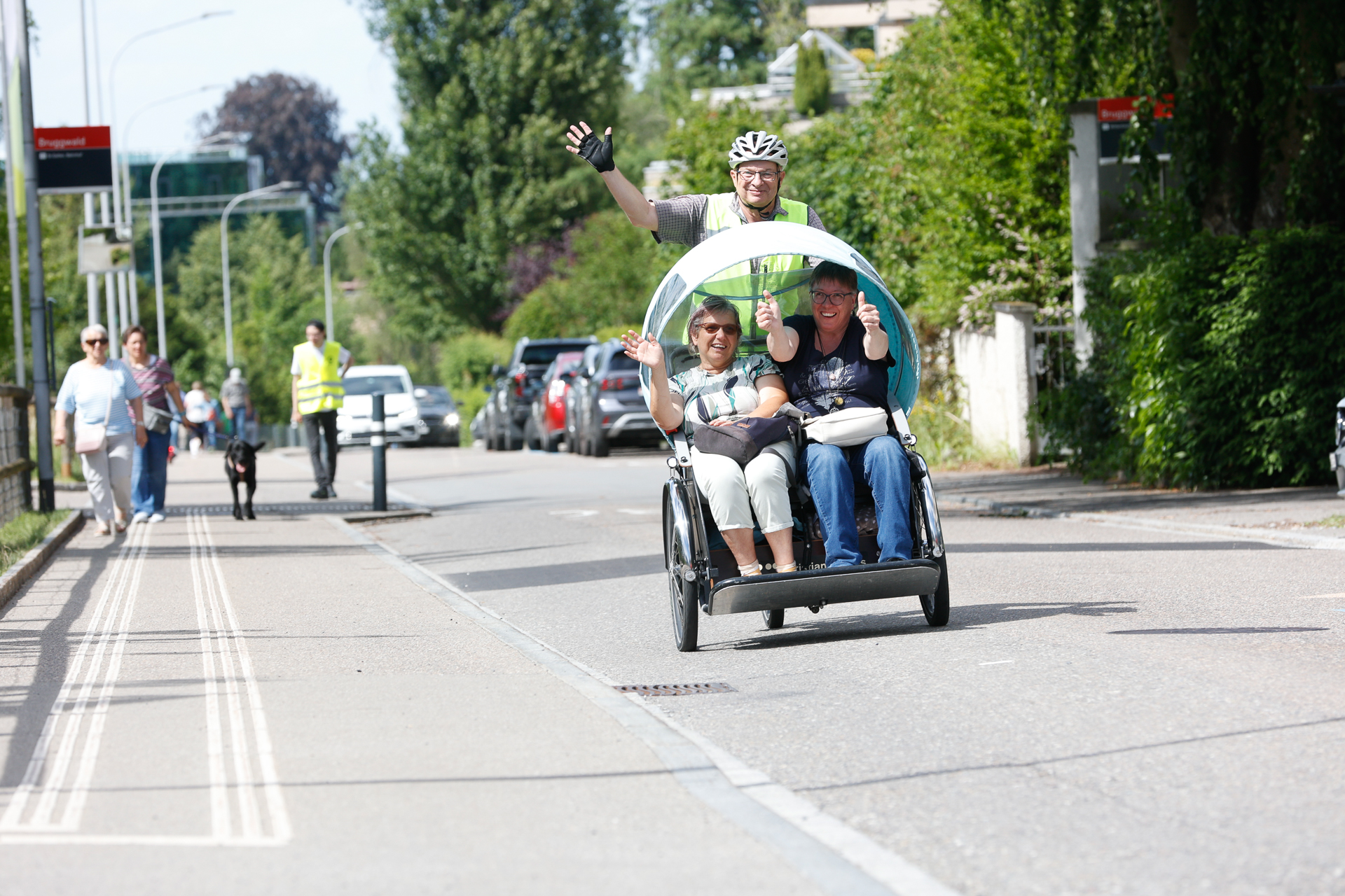 Ein Radfahrer mit Helm treibt eine Rikscha mit Verdeck an, in der zwei lächelnde Passagiere sitzen, die winken. Die Fahrt führt über eine Stadtstraße, die von Bäumen, geparkten Autos und Fußgängern gesäumt ist.