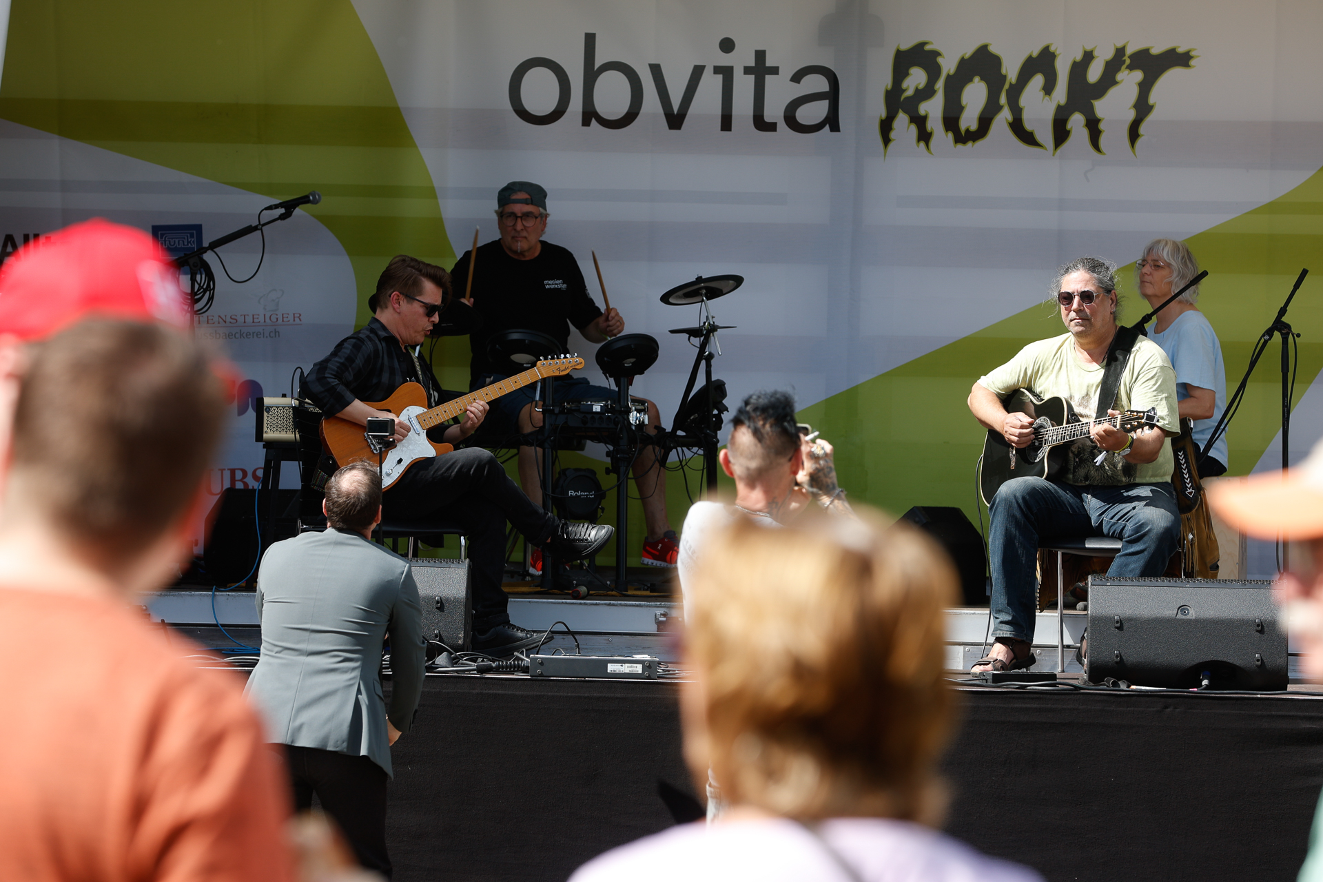 Eine Band tritt auf einer Open-Air-Bühne auf, auf der zwei Gitarristen sitzen und ein Schlagzeuger spielt, während das Publikum zusieht. Auf dem Hintergrund steht in fetten Buchstaben „obvita ROCKT“.