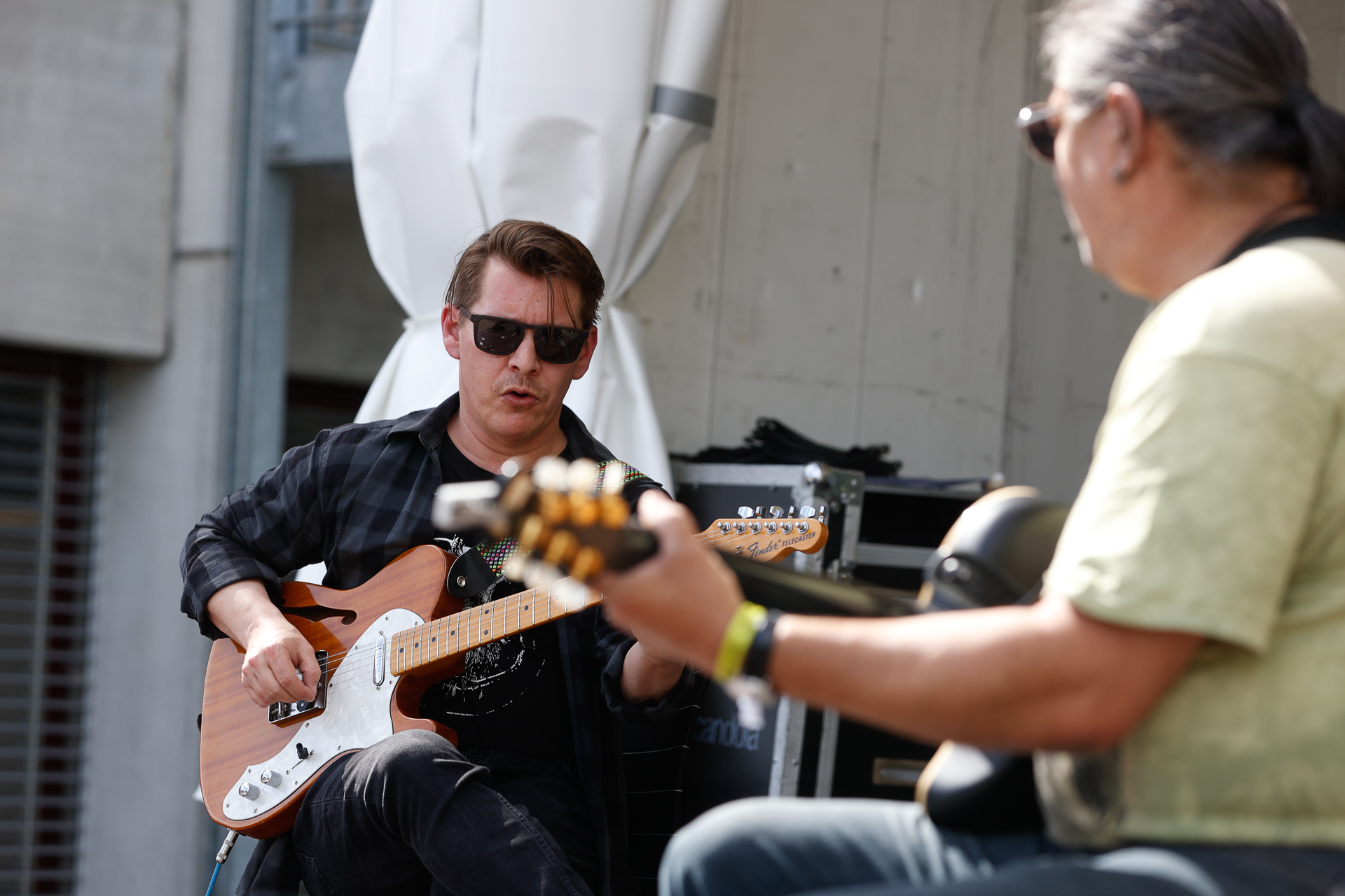 Zwei Musiker spielen im Freien E-Gitarre; einer im Vordergrund trägt eine Sonnenbrille und ein kariertes Hemd, der andere ist leicht unscharf und trägt ein gelbes Hemd. Im Hintergrund sind Ausrüstung und ein Gebäude zu sehen.