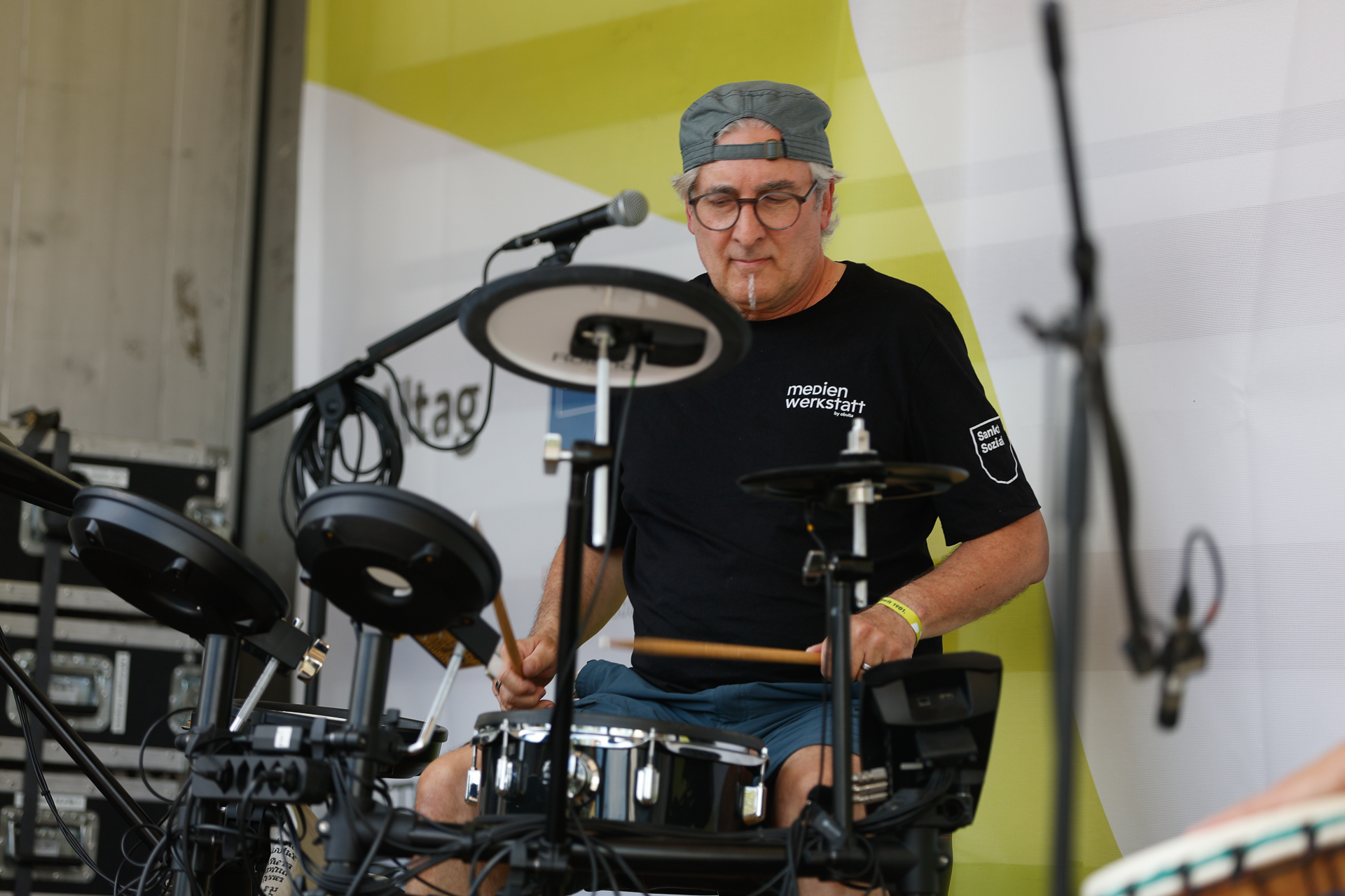 Ein Mann mit Brille, einer nach hinten gedrehten Kappe und einem schwarzen T-Shirt spielt auf einer Außenbühne auf einem elektronischen Schlagzeug und ist ganz auf seinen Auftritt konzentriert. Um ihn herum sind Tonanlagen und Mikrofone zu sehen.