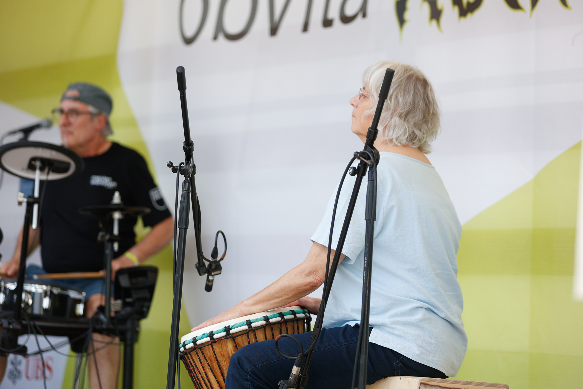 Eine ältere Frau spielt auf der Bühne eine Djembe, während im Hintergrund ein Mann ein elektronisches Schlagzeug spielt. Sie sind von Mikrofonen umgeben, und hinter ihnen hängt ein teilweise sichtbares Schild.