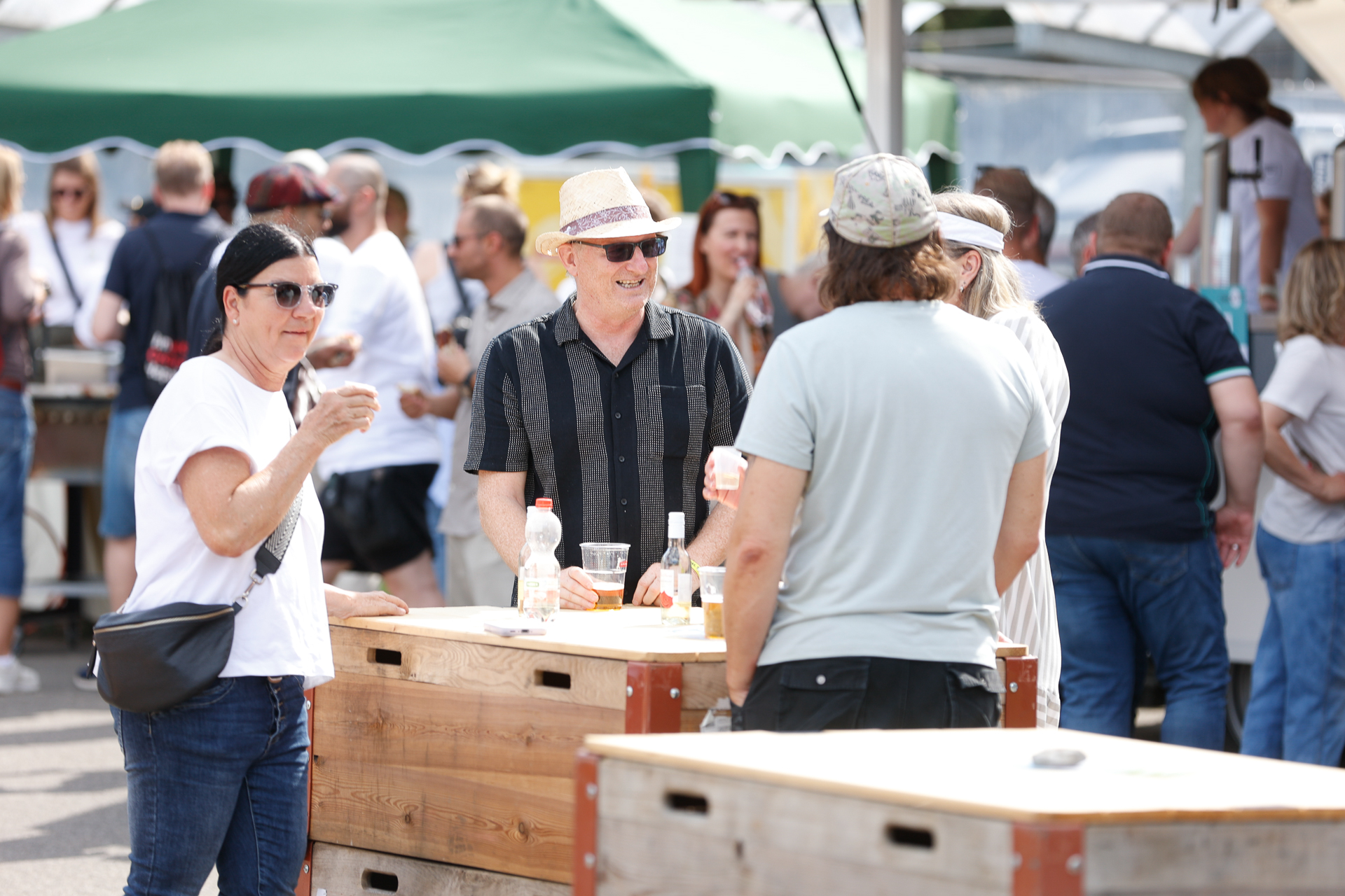 Eine Gruppe von Menschen unterhält sich und genießt an Holztischen bei einer Veranstaltung im Freien oder einem Festival ein paar Getränke, während sich im Hintergrund an einem sonnigen Tag andere unter die Gäste mischen und Zelte zu sehen sind.